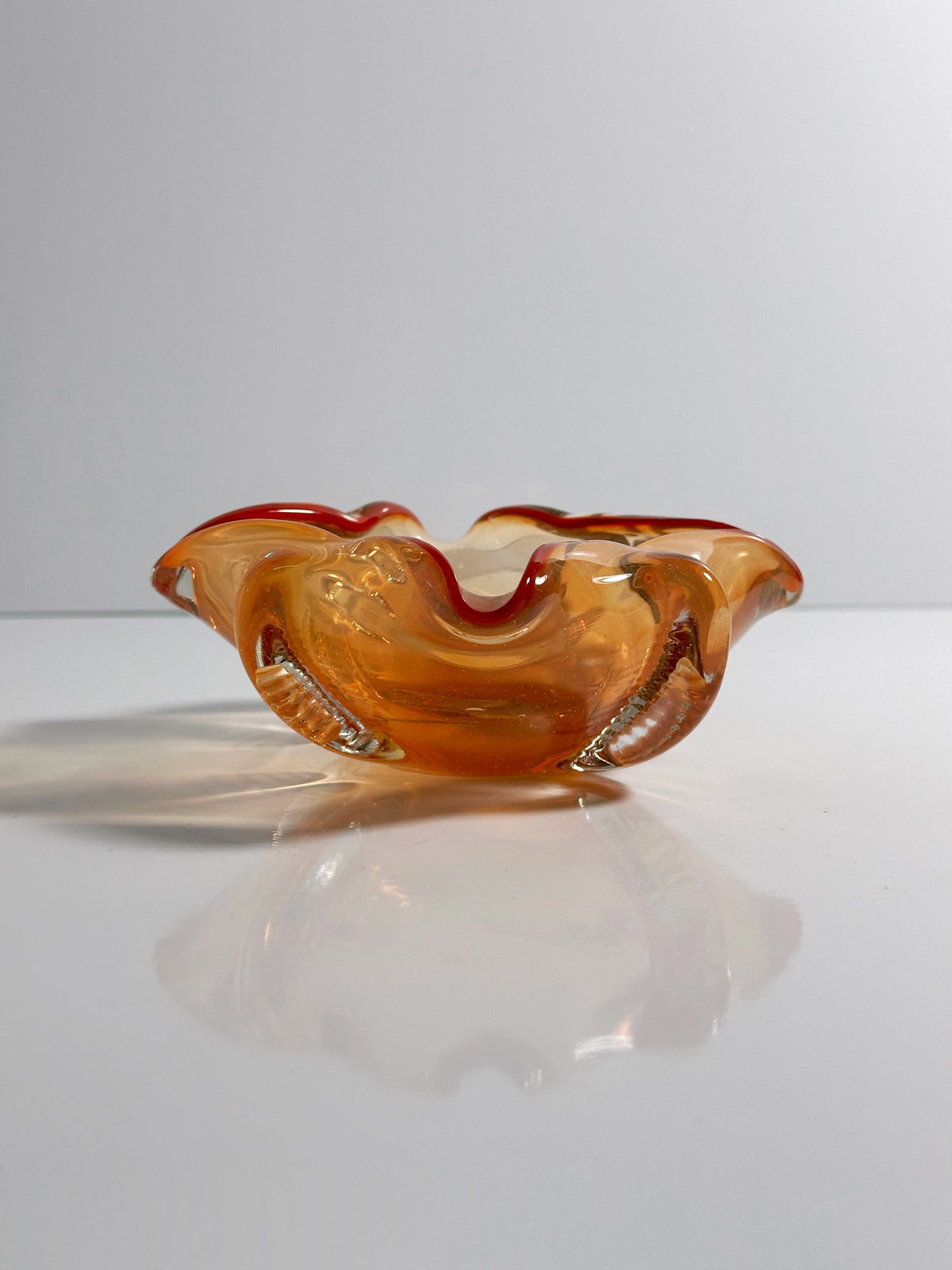 A translucent, wavy-edged Venetian Ashtray by Seguso Murano from the 1950s in amber and coral hues sits on a reflective white surface with a gray backdrop. Light reflections cast a subtle shadow beneath, evoking a nautical design element. Available at Dodo Vintage.