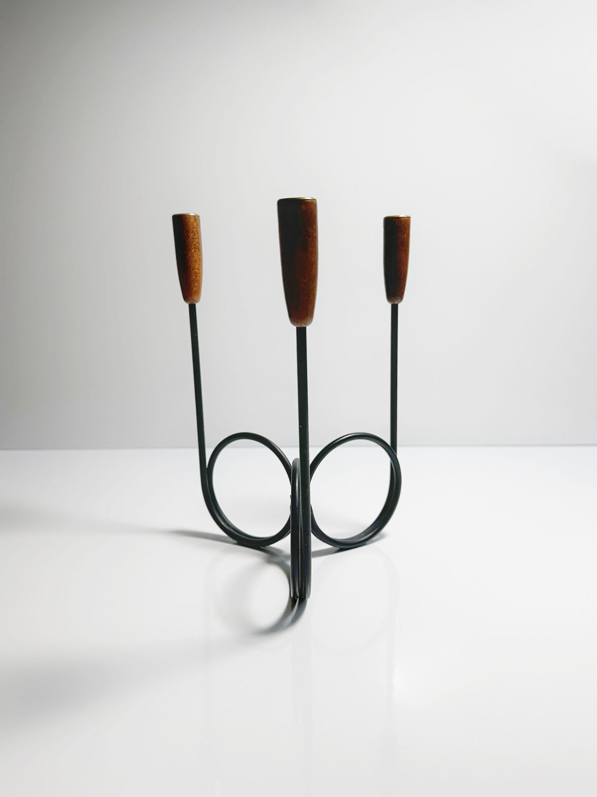 The Danish Candleholder 1960s by Dodo Vintage is a minimalist, modern three-armed candlestick holder featuring teak ends. Crafted from slender black metal rods that elegantly curve into a circular base, this stylish candelabra stands on a glossy white surface set against a plain light gray background.