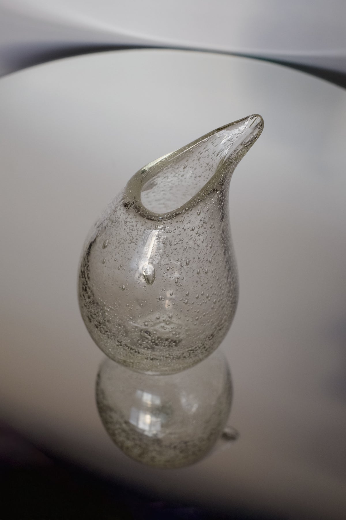 The Small Bubble Vase by septembre studios is a handcrafted clear glass sculpture shaped like a droplet with a pointed tip, standing on a reflective surface. Featuring small bubbles, it showcases exquisite craftsmanship that adds texture and depth to this art piece.