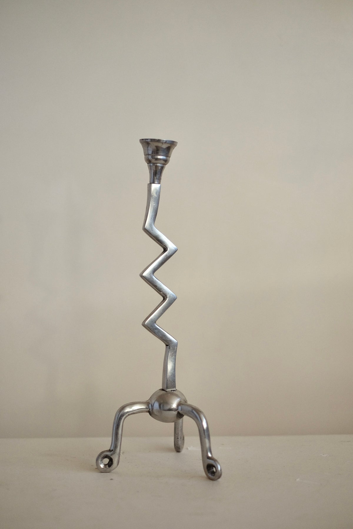 A stunning piece from the Set of Zigzag Candleholders by septembre studios stands proudly on a light surface against a neutral background. This metallic, zigzag-shaped holder boasts a shiny finish and is supported by three short, curved legs, making it a standout among candle holders.