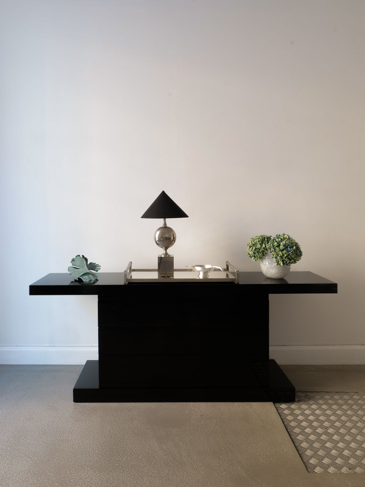 A High Gloss Console Table 1970s by Collection apart stands against a white wall. It holds a lamp with a triangular lampshade, a decorative leaf sculpture, a reflective tray with various items, and a round vase filled with green hydrangeas. The light gray floor features a textured mat.