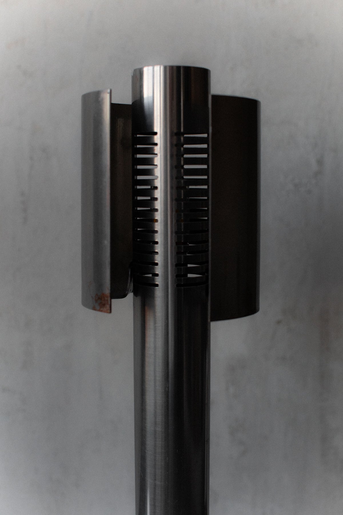 A close-up of the 1960's Space Age Lamps by Out For Lunch showcases a tall, sleek, cylindrical design with vertical slits and curved side panels against a muted gray background, highlighting its polished texture and timeless appeal.