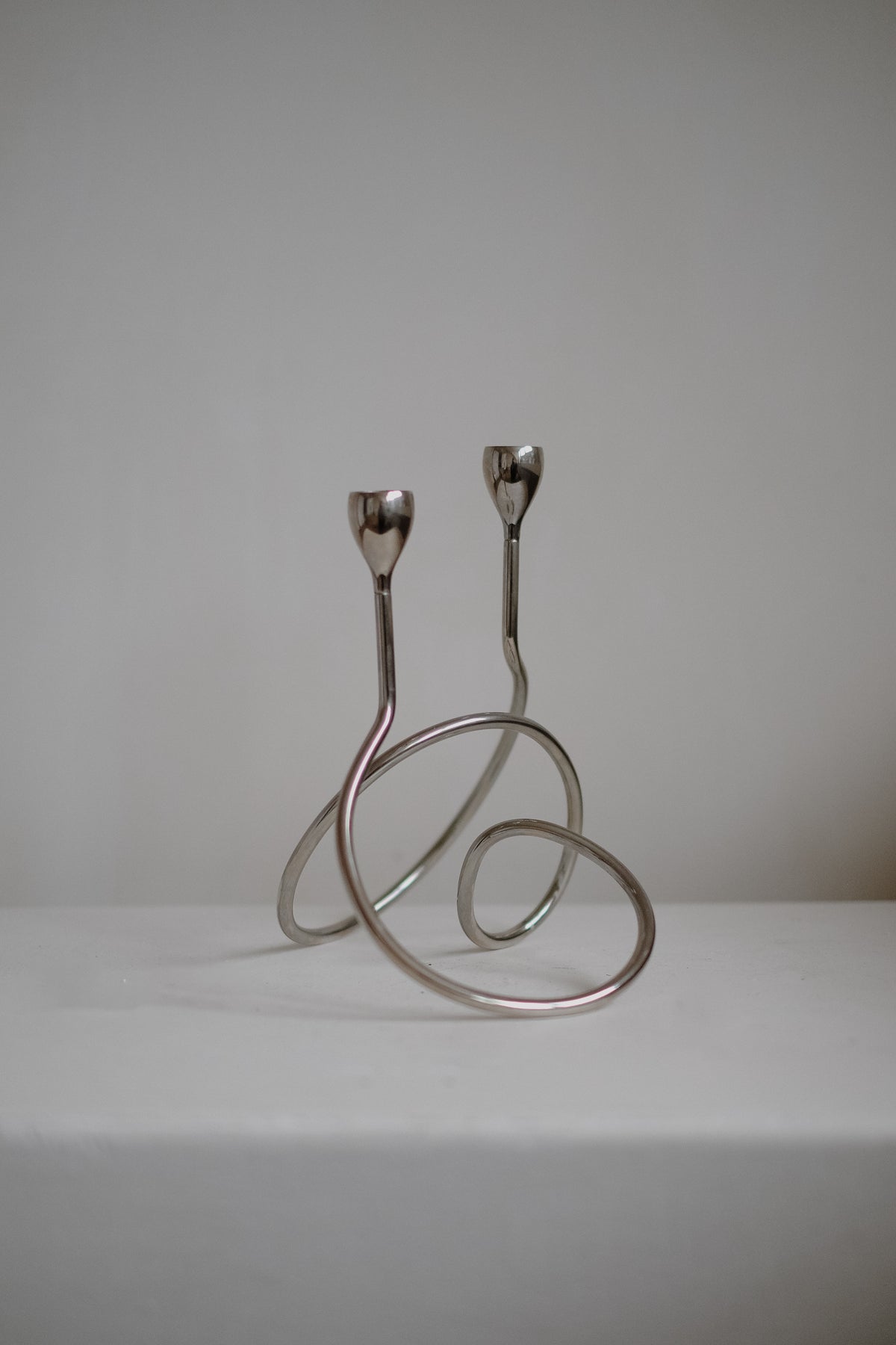 The "Loop" Candle Holder by septembre studios is a minimalist, abstract metal sculpture with chrome loops featuring two upward extensions akin to stems. This dynamic piece doubles as a functional dinner candle holder, blending art and utility against a plain backdrop.