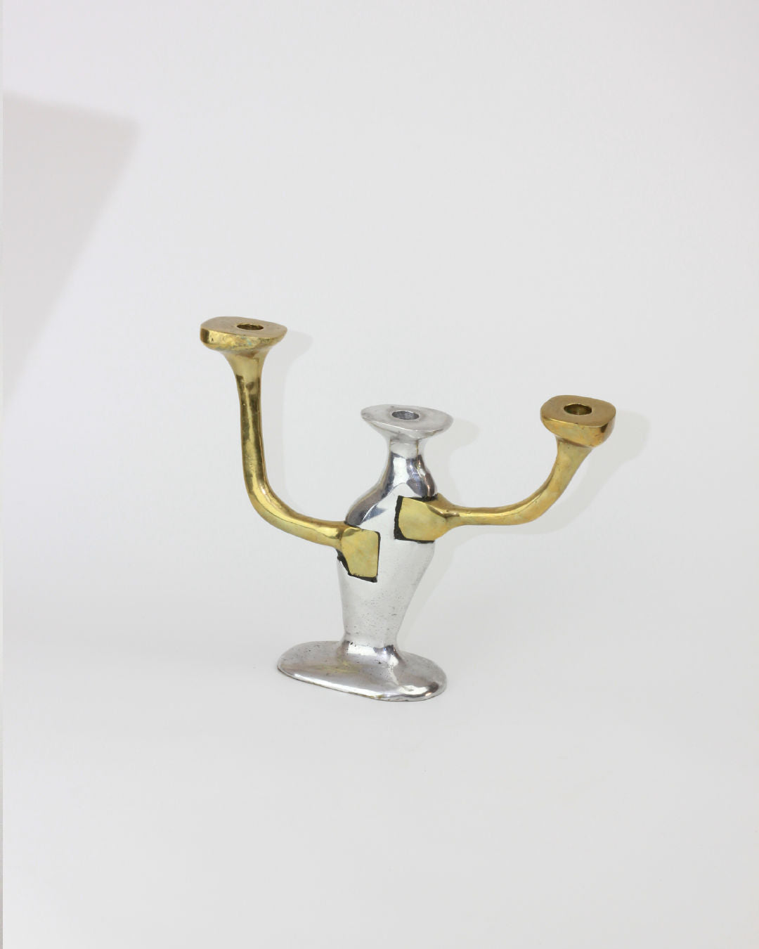 The Brutalist Candle Holder from Boga Avante Shop features two arms and a central support, artfully displayed on a white background. The left arm and central support boast a sleek silver finish, while the right arm adds contrast with its gold hue. Each arm has a circular opening at the top for holding candles, beautifully capturing the essence of 1970s Seville design.