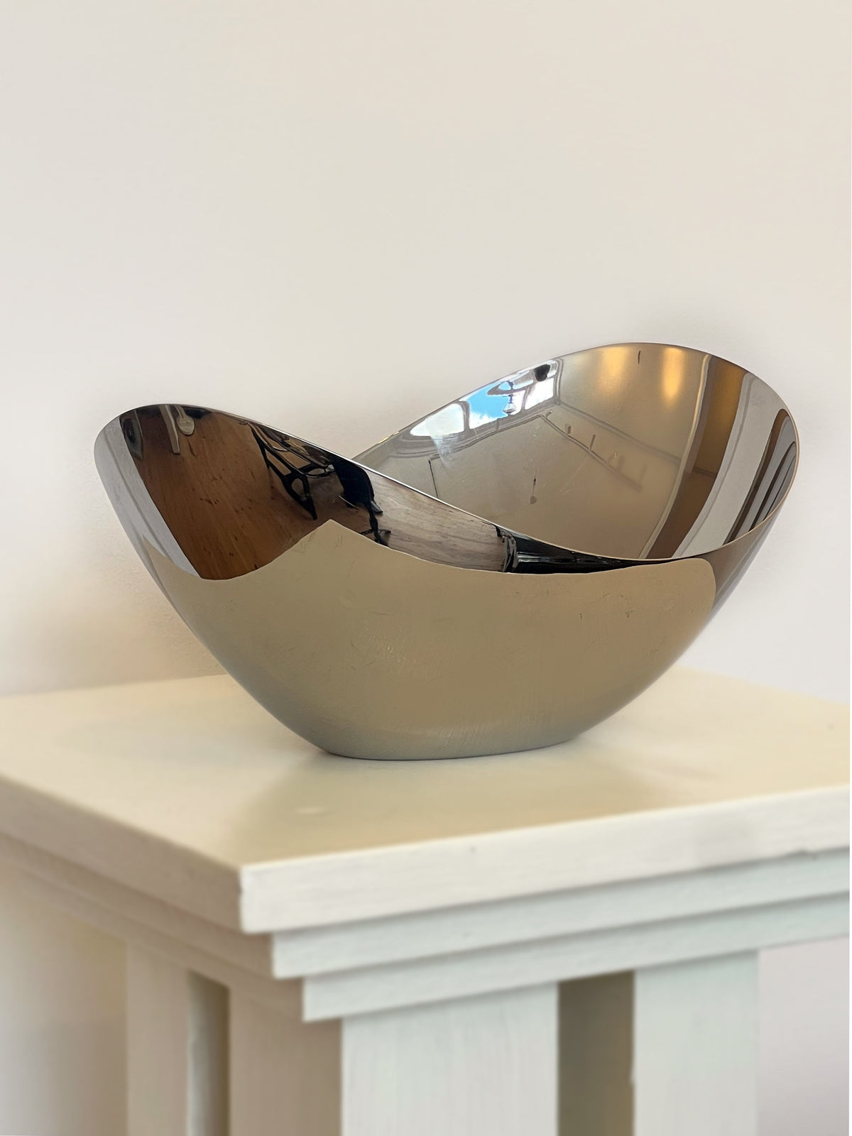 A shiny, reflective Porter Studio George Jensen Bloom Bowl with curved edges sits on a light pedestal against a white wall. Made from stainless steel, the bowl’s surface perfectly mirrors its surroundings.