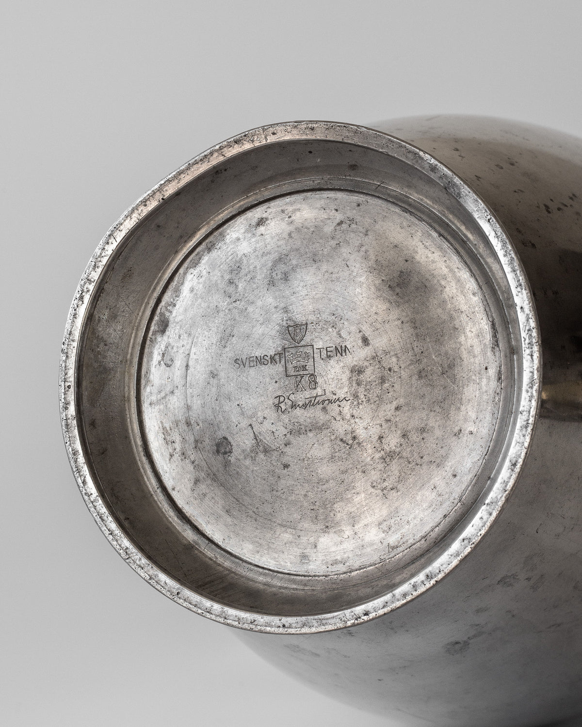 The image depicts the underside of a Large Modernist Pewter Amphora from the 1930s by Spigel, with engraved "Svenskt Tenn" and initials, featuring a worn surface and patina similar to Rolf Engströmer's style.
