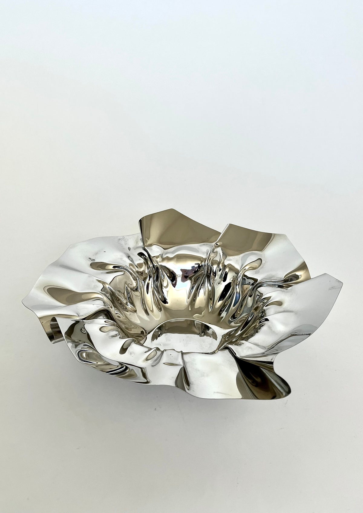 The Folded Chrome Bowl by septembre studios features a shiny, reflective surface with a wide, wavy edge and a deep center. Against a plain white background, its intricately textured ripples and folded shapes create a modern and artistic appearance—perfect for enhancing any interior setting.