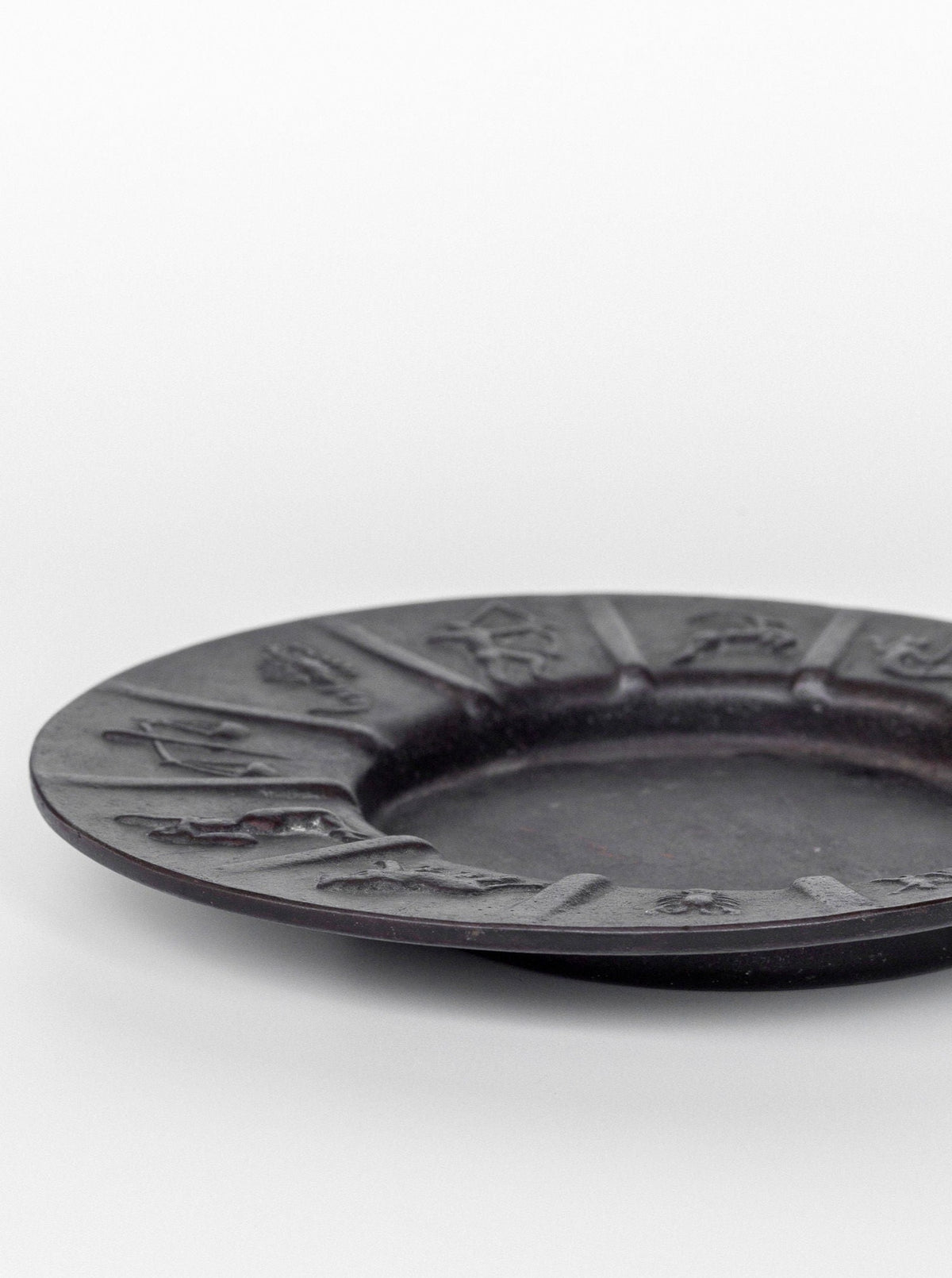 The Spigel Art Deco Zodiac Signs Decorative Dish is a dark, circular plate with raised, ornate designs around its outer edge, featuring different symbols or figures evocative of a zodiac theme. The vintage iron dish has a flat center and is photographed on a plain white background.