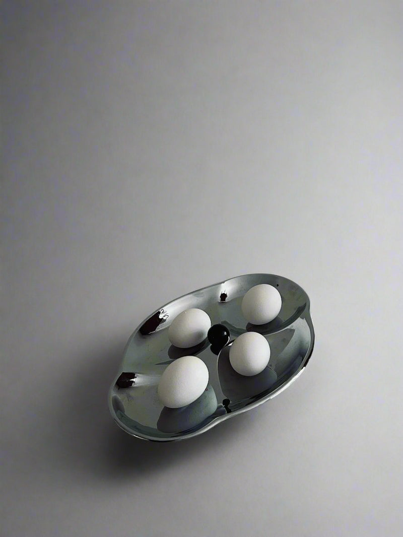 A sleek, modern Mid Century Serving Tray by Flock Studio, featuring an abstract design and a silver finish, holds four white eggs. Its reflective surface stands out against the plain, muted background, creating a minimalist and artistic composition reminiscent of Mid Century aesthetics.