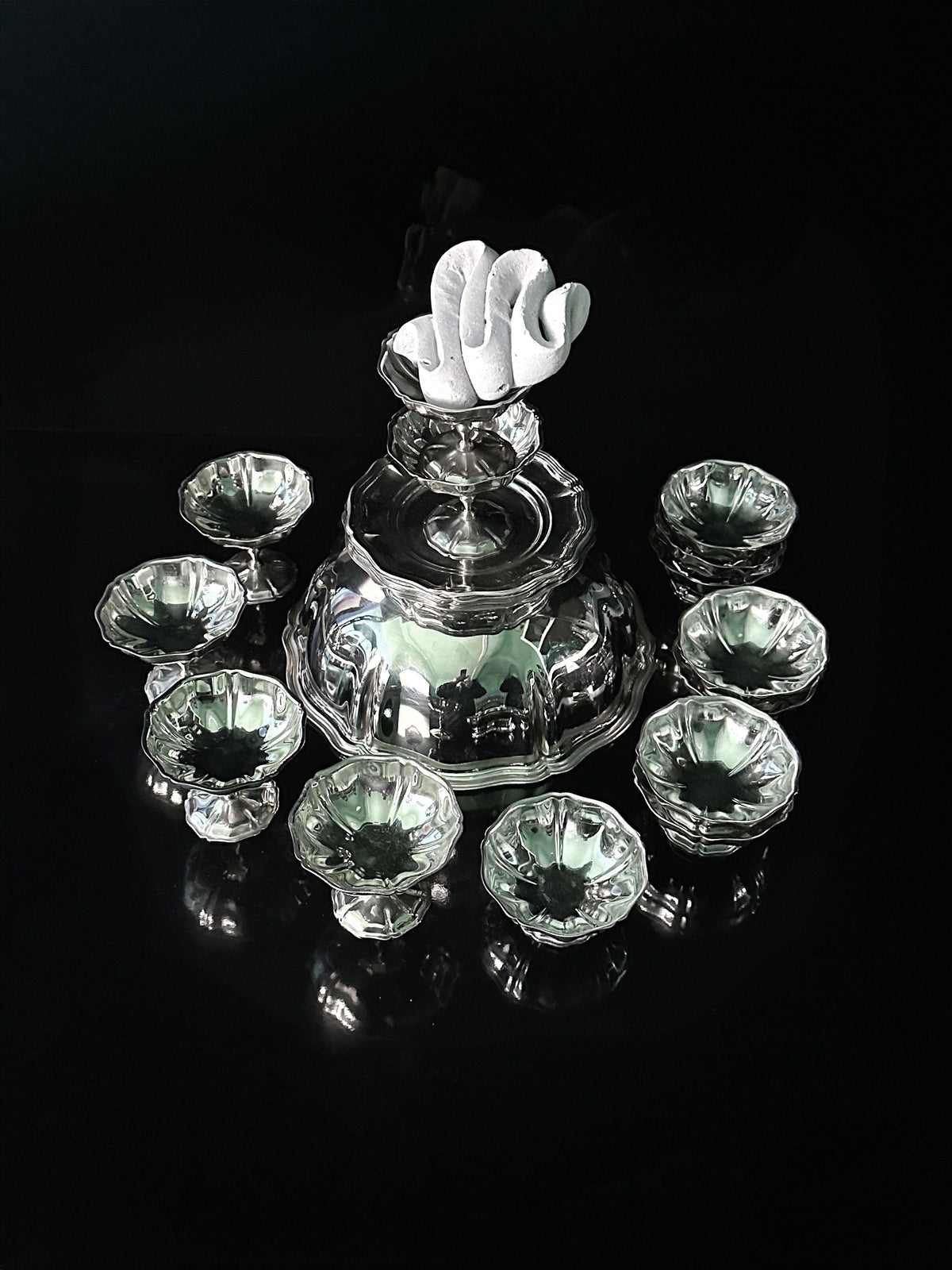 The Dodo Vintage Italian Stainless Steel Dessert Coupe Set features a larger covered bowl with decorative white fabric, centered on a glossy black surface, and surrounded by eight smaller matching bowls that reflect light beautifully.