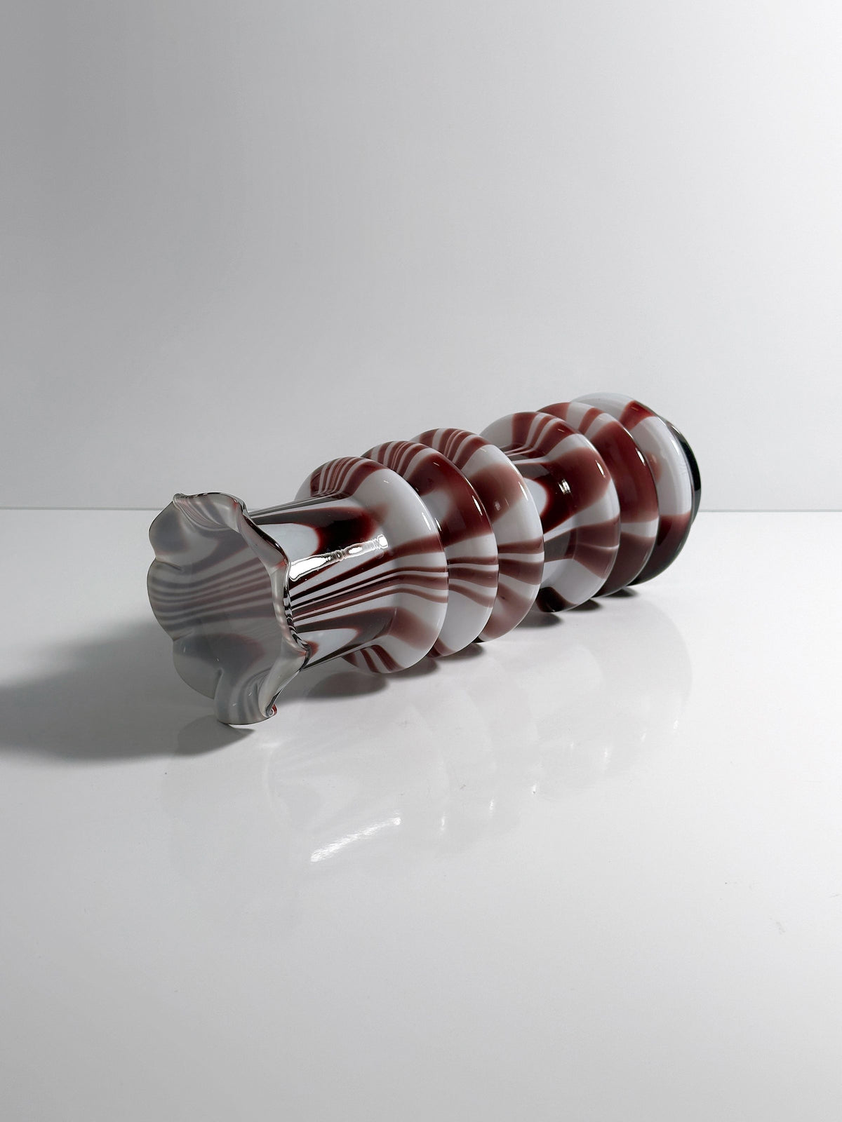 A 1970s Carlo Moretti Opalina Fiorentina vase from Dodo Vintage, showcasing a spiral design with white and maroon stripes, lies horizontally on a reflective surface. The opal glass colors create a vibrant mirror image against the plain white background.