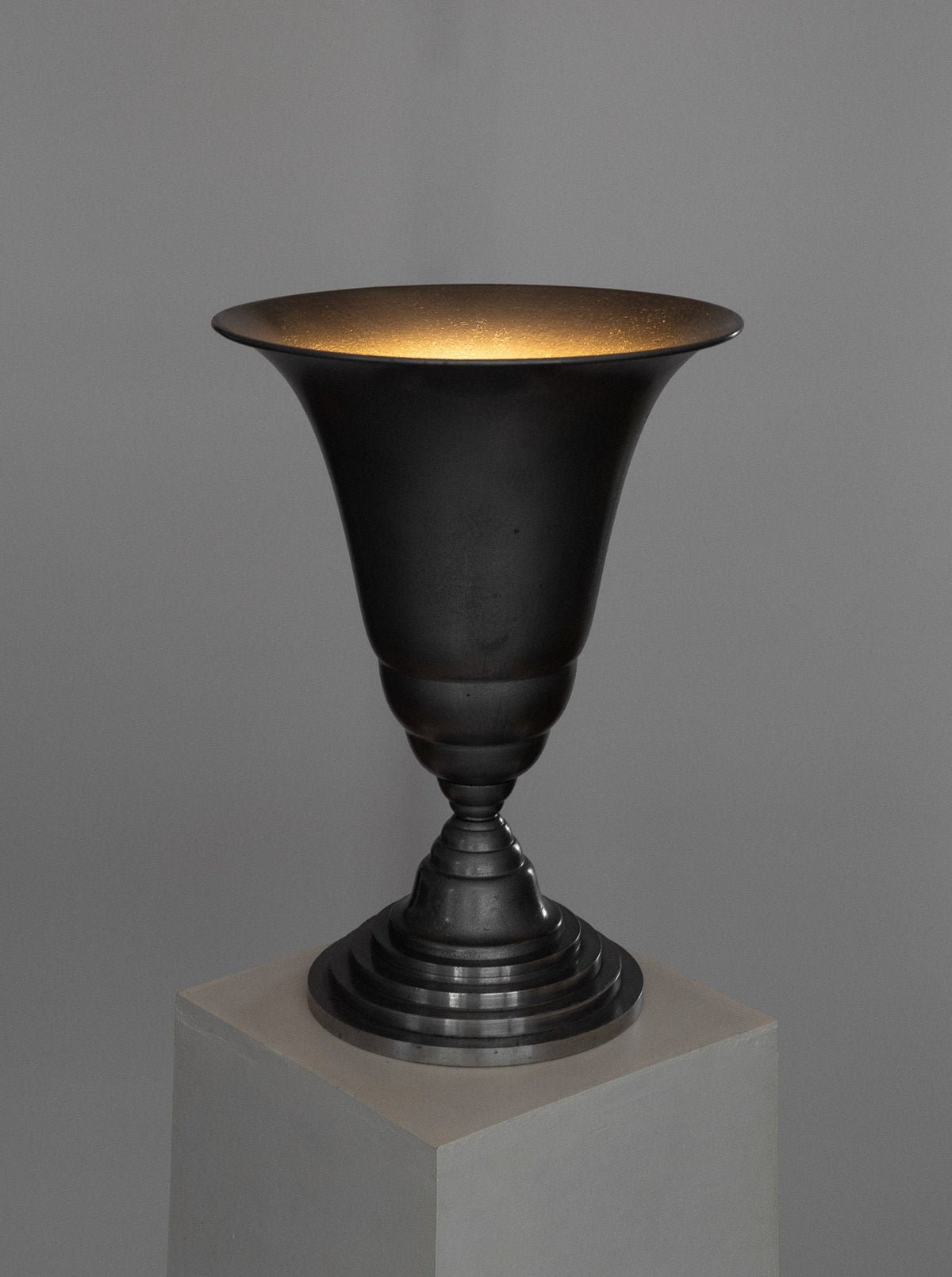 A dark, metallic Art Deco Torchière Table Lamp from France with a wide-brimmed and smooth, concave shape along with a textured base, displayed on a simple white pedestal against a gray background. The lighting creates a soft glow inside the lamp reminiscent of French Art Deco lighting, highlighting its polished surface and elegant design under the renowned brand Spigel.