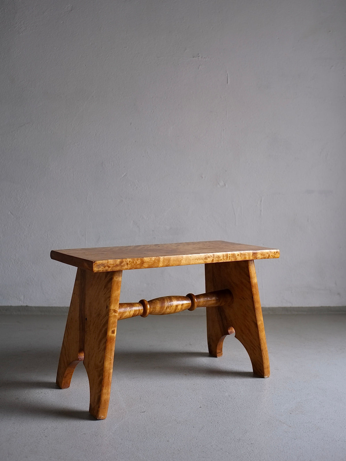 A simple Rustic Solid Karelian Birch Stool | Latvia | 1950s in vintage condition with a light, natural finish positioned against a plain gray wall on a concrete floor. Crafted from solid Karelian birch wood by Veter Vintage, the stool has arched legs connected by a horizontal rounded bar and an overall rustic appearance.