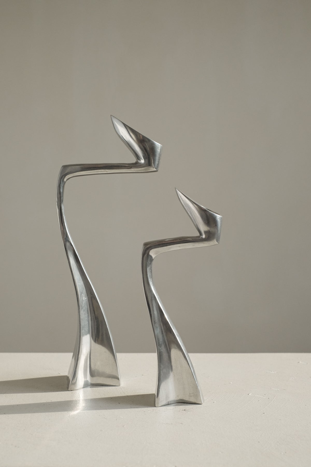 The Pair of Candleholders by Matthew Hilton 80's, from septembre studios, features two abstract, silver metallic sculptures with sleek, curved forms. They display elongated shapes resembling figures facing each other on a light surface against a plain background, captivating design enthusiasts.