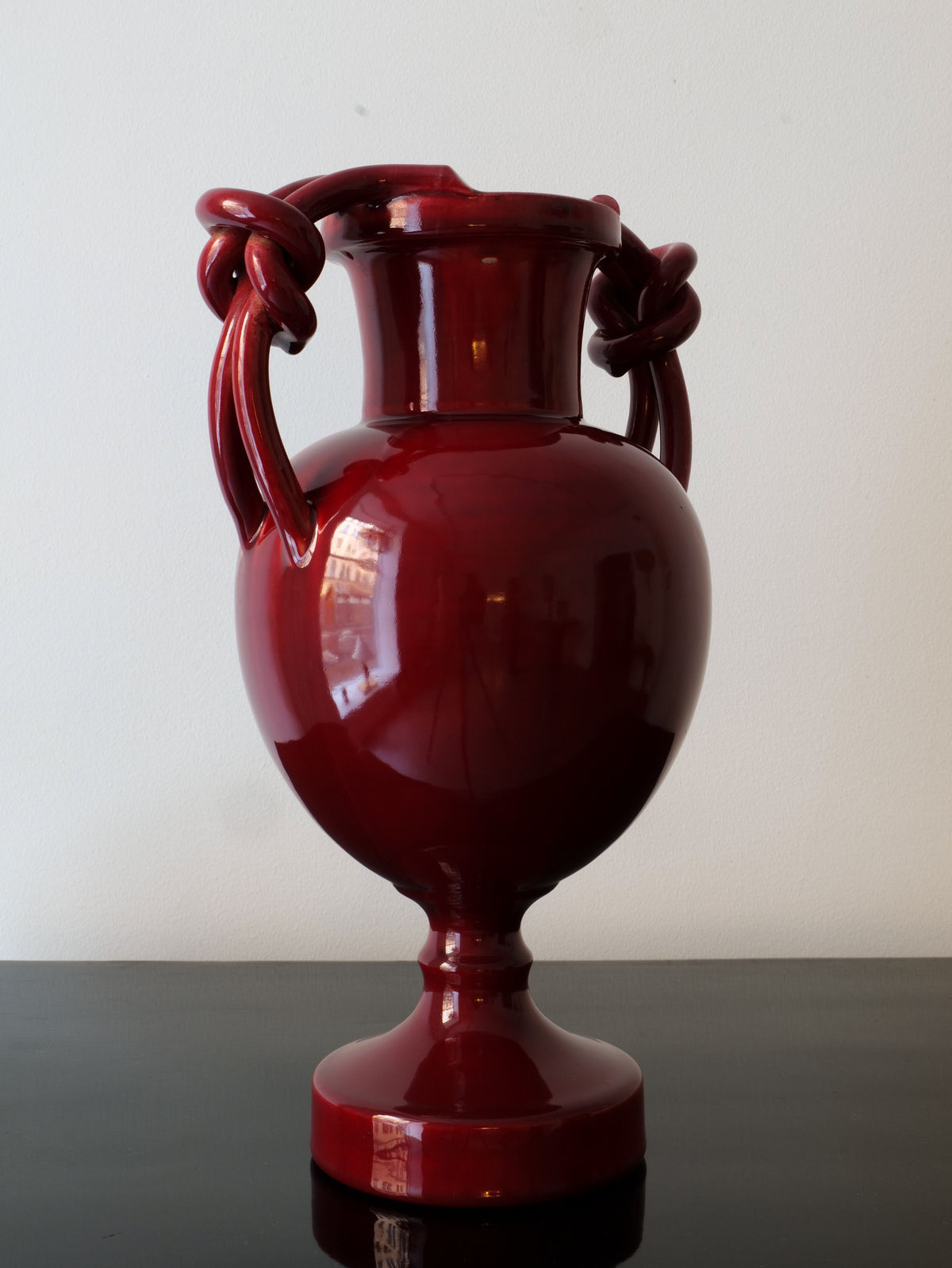 A glossy, dark red ceramic Baluster Vase Jérôme Massier by Collection apart, reminiscent of Vallauris ceramics, features two intricately knotted handles and rests on a polished black surface. The vase's reflective finish captures the light, emphasizing its smooth and elegant form against a plain white background.