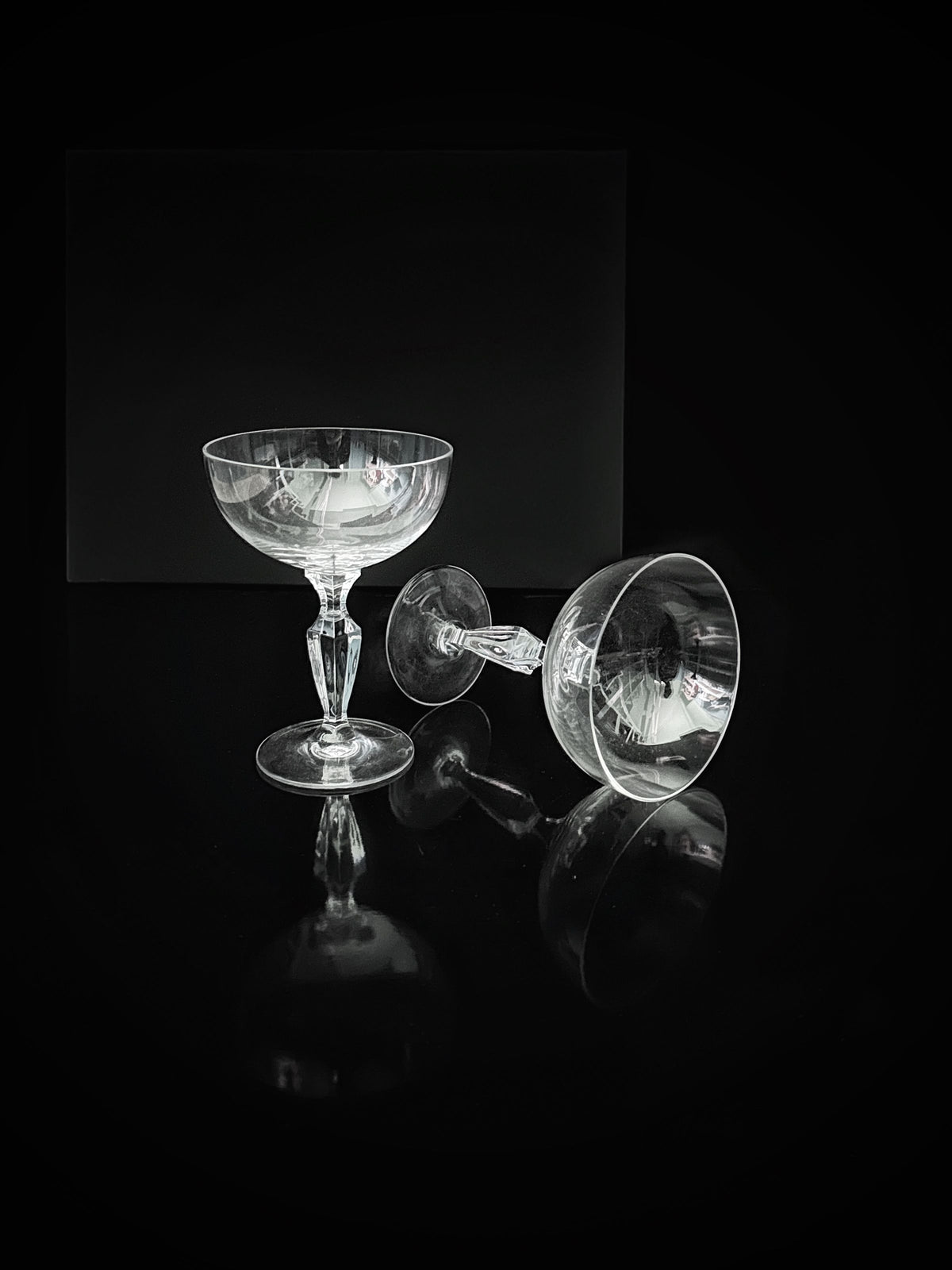 Two Dodo Vintage Mid Century Crystal Champagne Coupes, exemplifying timeless craftsmanship, are elegantly positioned against a glossy black backdrop. One coupe stands upright while the other reclines gracefully on its side, with the reflective surface enhancing their sophisticated arrangement.
