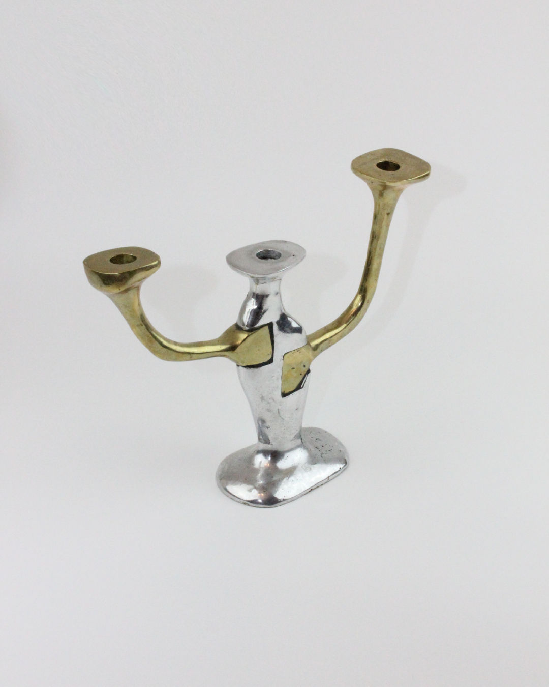 Presenting the Brutalist Candle Holder by Boga Avante Shop: with a unique design featuring a central base and two upward-curving arms. Crafted in silver for the base and central structure, and gold-colored arms to mimic brass and aluminum, this holder can accommodate two candles.