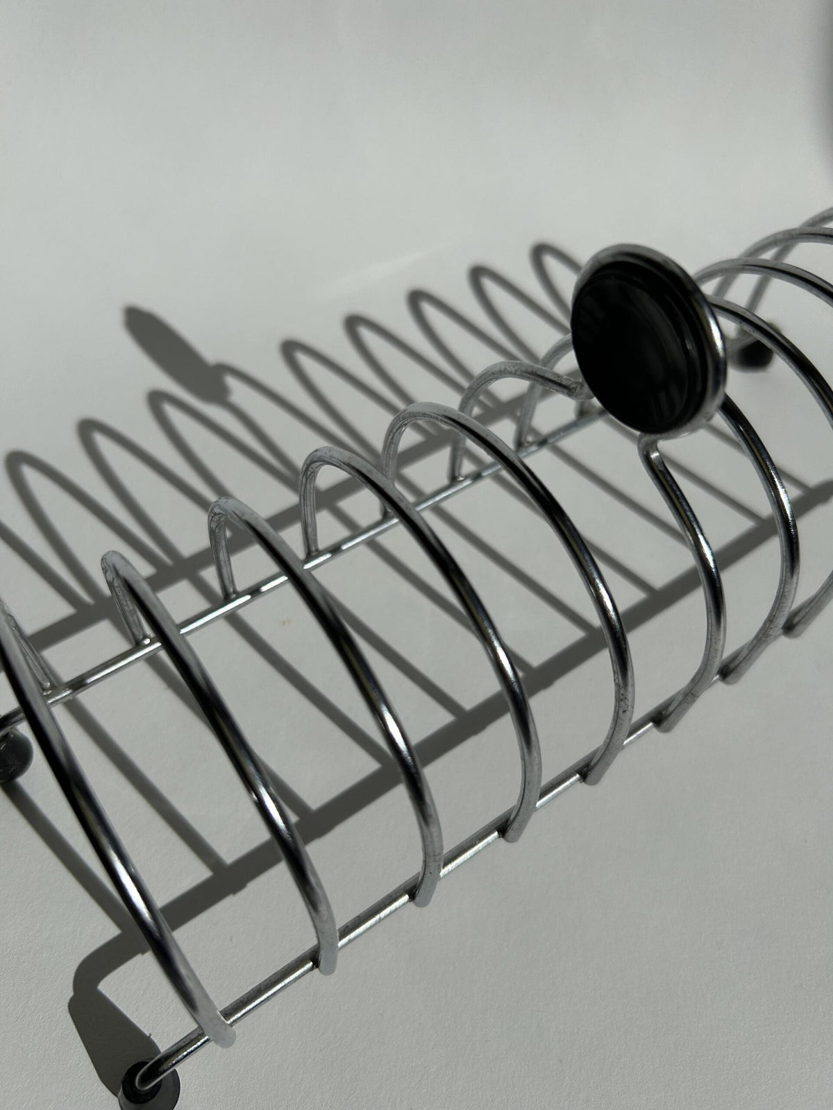 A Postmodern Toast Rack from Flock Studio, inspired by Tomado Holland, features a silver metal spiral design reminiscent of the 1950s. It rests on a white surface casting a shadow, with a black cap accentuating its end and echoing elegant postmodern style.