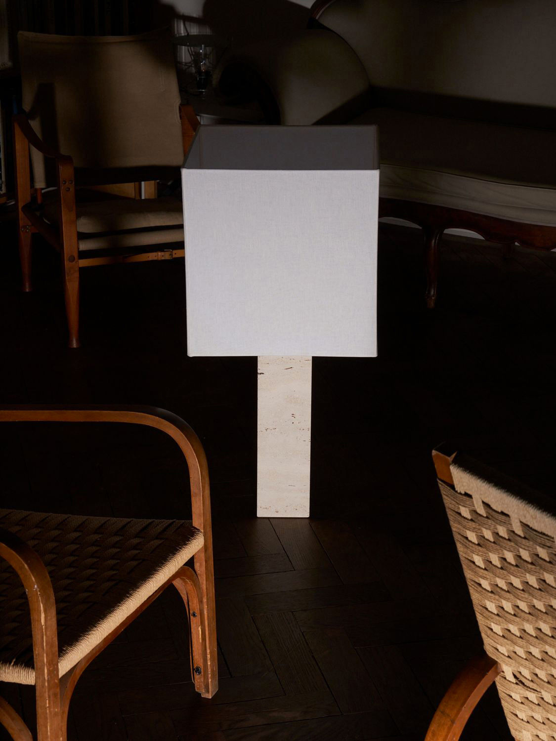 A minimalist room features two wooden chairs with woven seats and armrests. A modern rectangular floor lamp, the Roma I Squared White Travertine by Nassi Lamps, with a pure linen shade stands between the chairs. The room has low lighting, creating a stark contrast between the illuminated lamp and the dark surroundings, highlighting its modernism.