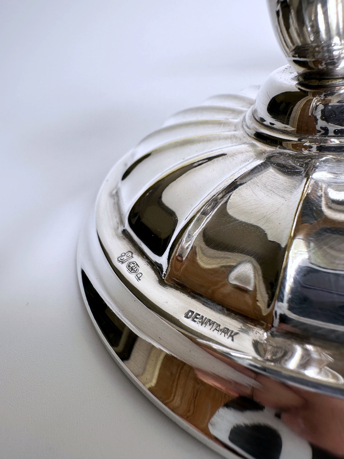 A close-up of a shiny, polished silver object, possibly the base of the Danish Mid Century Candleholder by Dodo Vintage. The object features ornate details and bears engravings including "DENMARK" and several hallmark stamps on its surface. The background is out of focus.