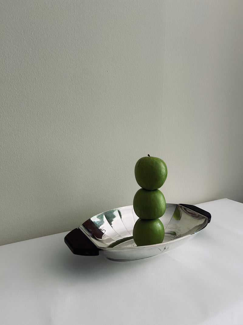 A minimalist image of three green apples stacked vertically on the Flock Studio Art Deco Serving Tray. The tray, crafted from polished metal with handles, rests on a white surface against a plain light-colored wall. Soft lighting casts subtle shadows, enhancing the modernist design's elegance.