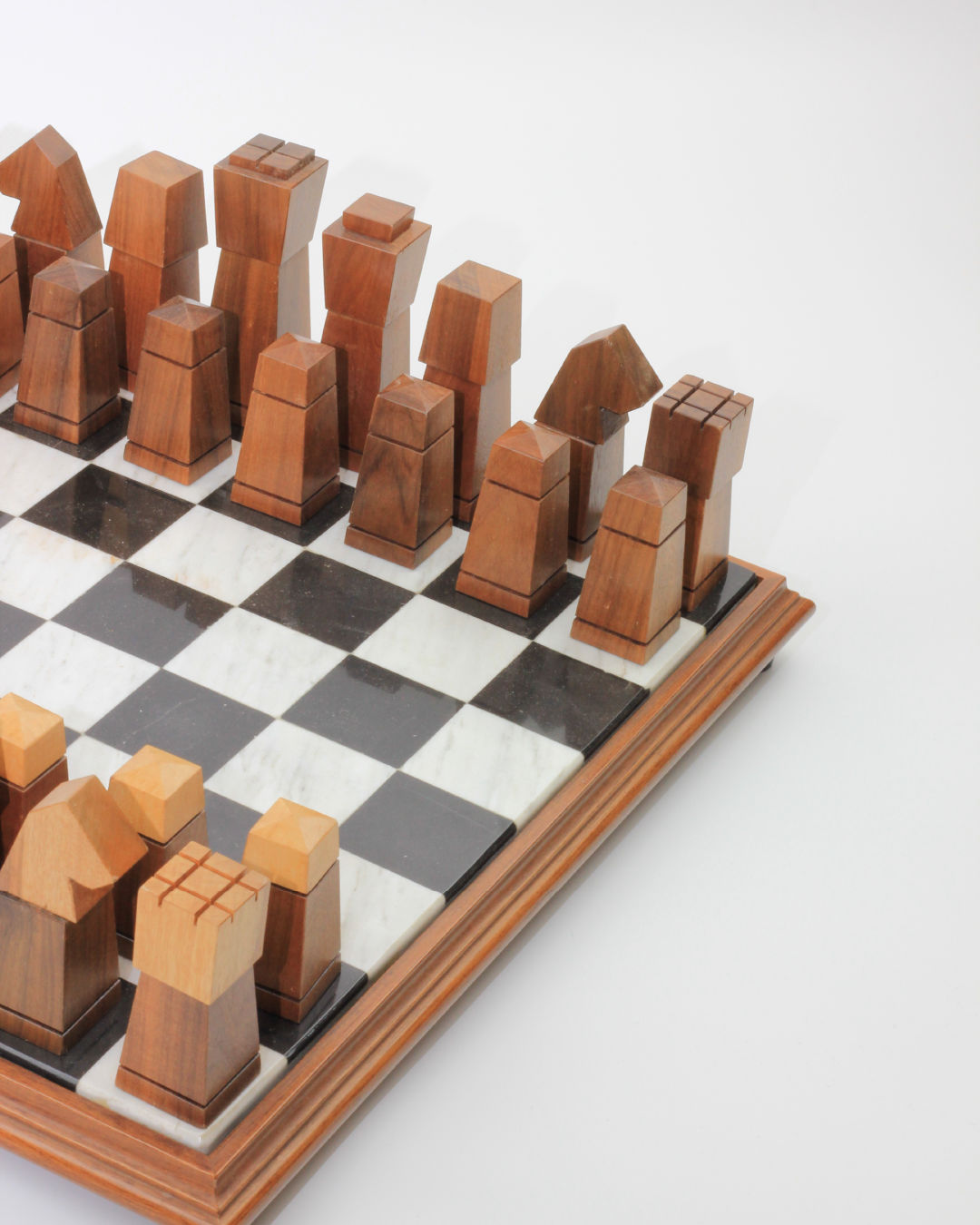 Introducing the Wooden and Marble Chessboard by Boga Avante Shop. This modern chess set features a stunning wooden and marble chessboard, with uniquely designed hand-carved pieces that boast geometric shapes, offering a contemporary twist on traditional game pieces. The scene highlights one side of the board with the pieces meticulously set up, ready for play.
