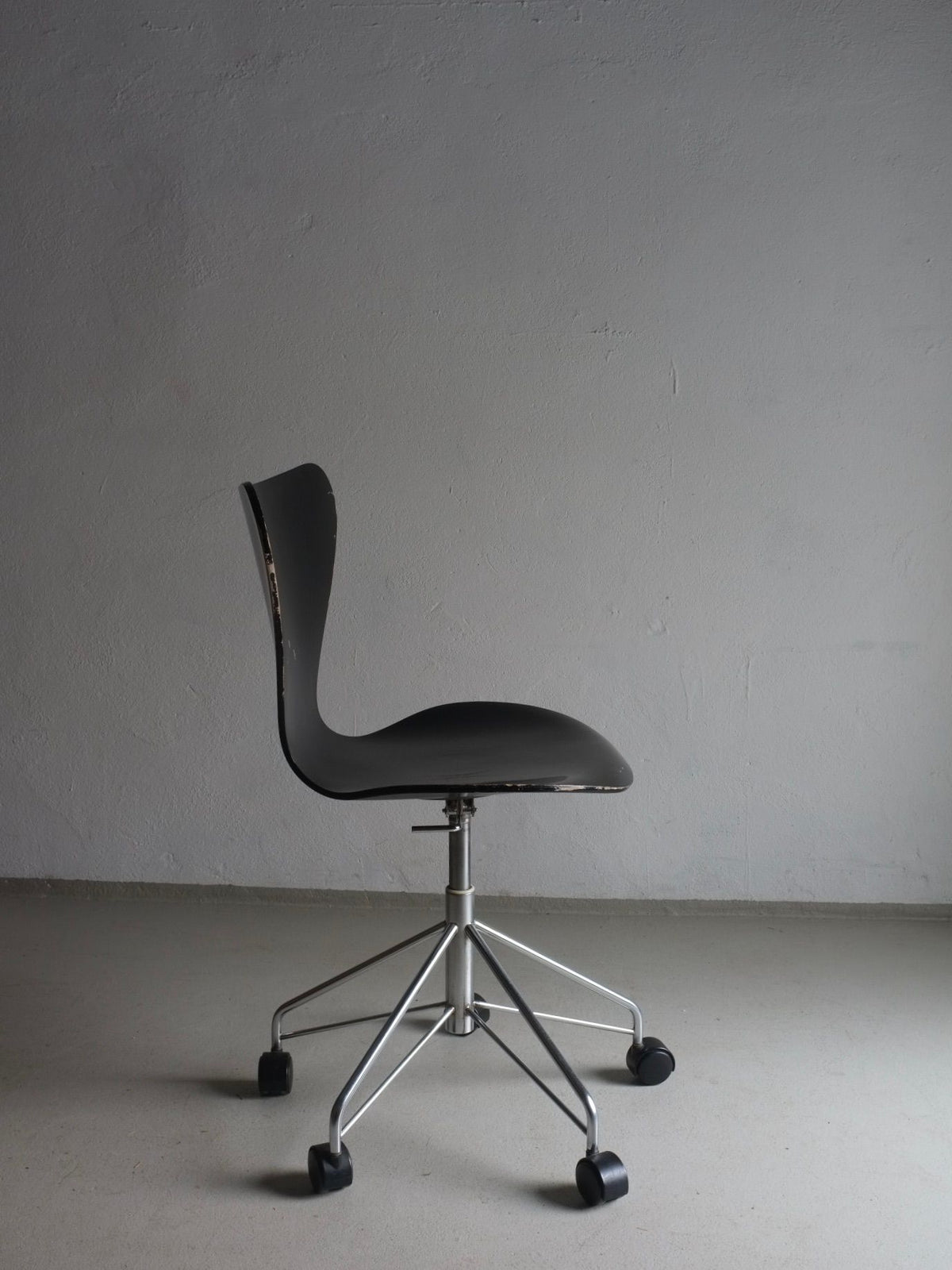 A modern office chair with a sleek black seat and backrest stands against a plain, light-colored wall. Reminiscent of Arne Jacobsen's design for Fritz Hansen, this 3117 Black Swivel Chair by Arne Jacobsen for Fritz Hansen from Veter Vintage features a minimalist metal base, five legs, and caster wheels. The setting appears to be a simple, undecorated room.