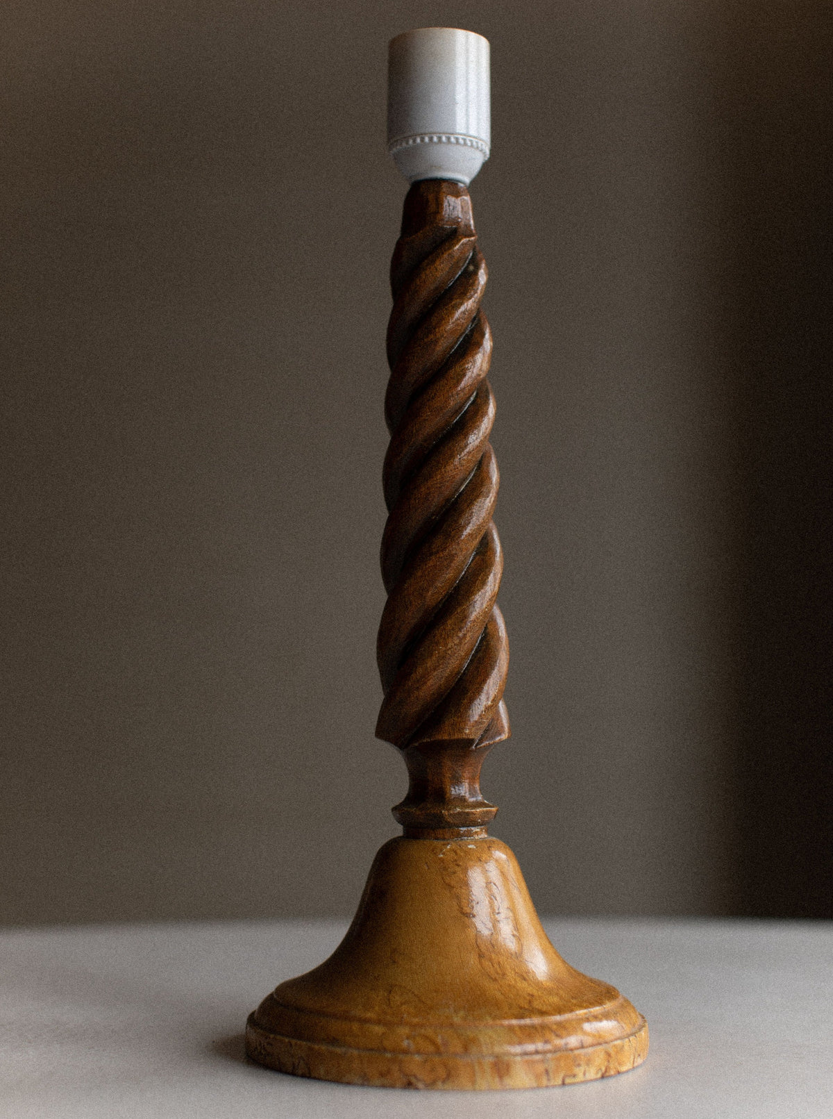 A Twisted Wooden Lamp from Out For Lunch stands on a flat surface, showcasing its natural wood grain. The warm, polished finish highlights the artisanal craftsmanship. The top is fitted with a white lightbulb socket, though no bulb is present. The background is neutral and slightly blurred.