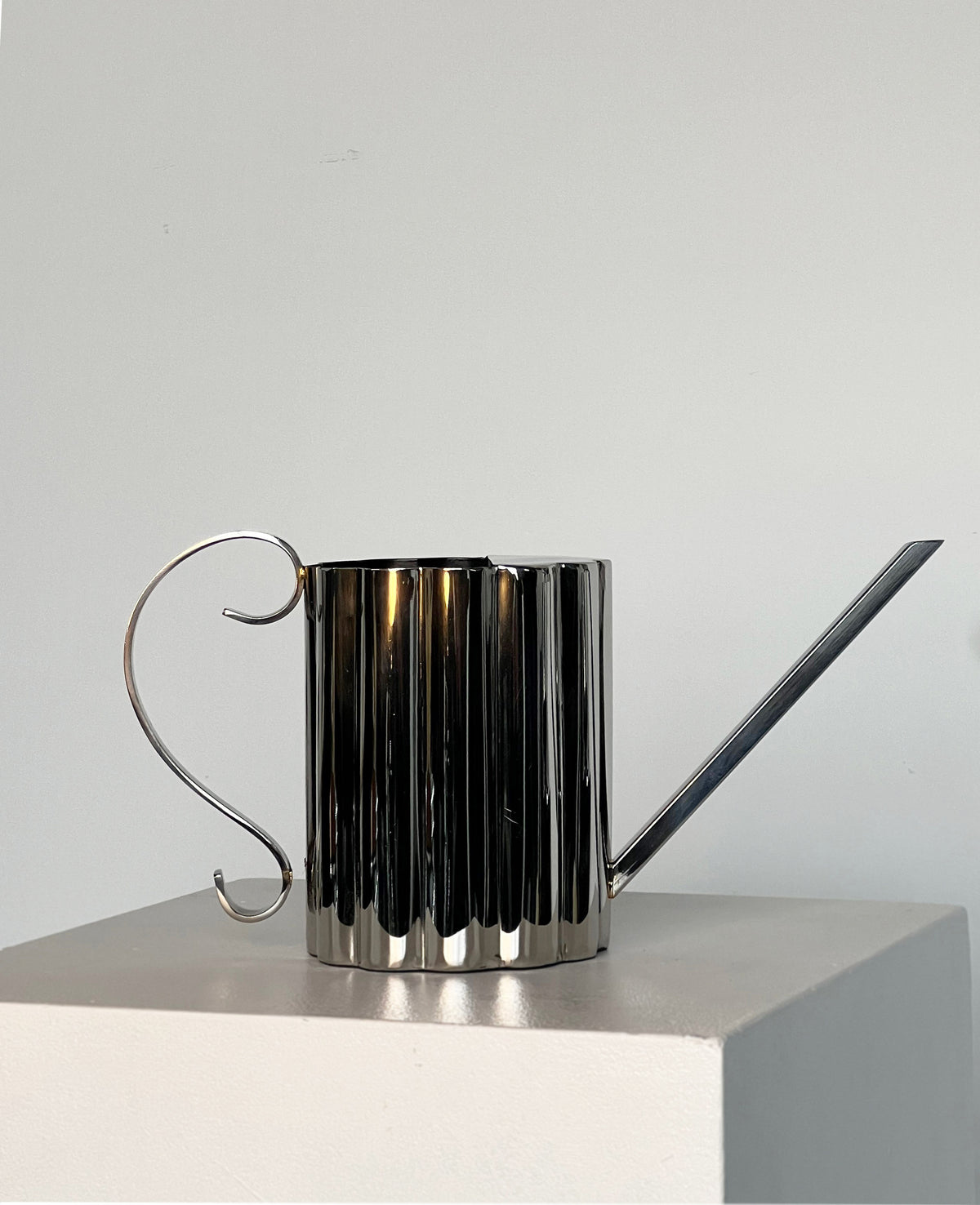 The Fluted Watering Can by Porter Studio, featuring a ribbed cylindrical body, elegantly curved handle, and long, narrow spout, creates an ornate silhouette on a white pedestal against the plain grey background.