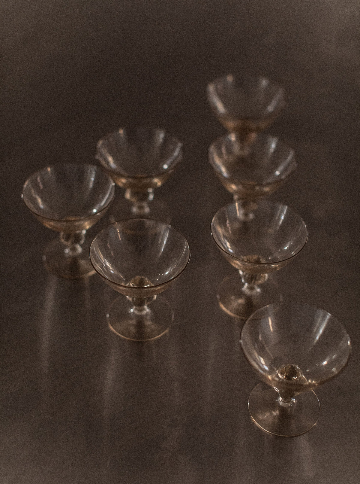 A photo of seven empty, clear glasses from the "Set of 7 Art Deco Glass" by Out For Lunch, arranged in a staggered pattern on a reflective, dark surface. The glasses' reflections are visible, and the image has a soft, warm-toned lighting that enhances the sophistication of the drinking experience.