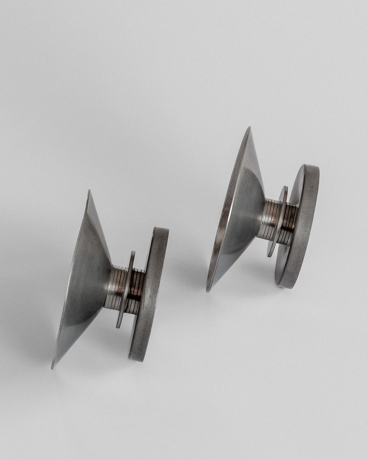 Two metallic, conical Modernist Candleholders by Sylvia Stave (Spigel, 1930s) stand upright. Their pointed ends face up with bases connecting cylindrical sections. Resembling vintage pewter candlesticks, they cast subtle shadows as they sit side by side.