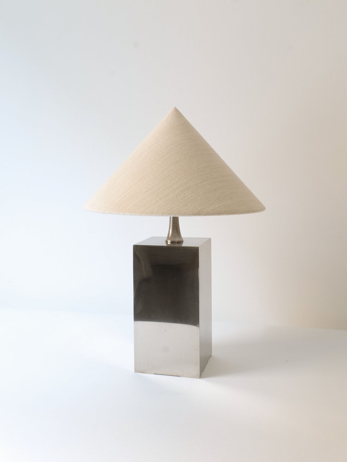 The Barbier Small Box Lamp 1970s by Collection apart features a minimalist design with a rectangular, chrome over brass base and a beige, cone-shaped fabric lampshade. The plain white wall background emphasizes the lamp's simple and elegant aesthetic, reminiscent of French 1970s lighting.