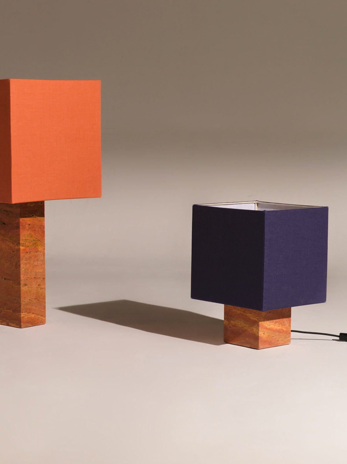 Two square-shaped, modernist Nassi Lamps Roma I Squared Red Travertine with blocky bases are shown against a gray background. One lamp has an orange shade and the other a navy blue shade, both crafted from pure linen. Their bases appear to be made of brownish, stone-like natural materials, and the cord of the navy lamp is visible.
