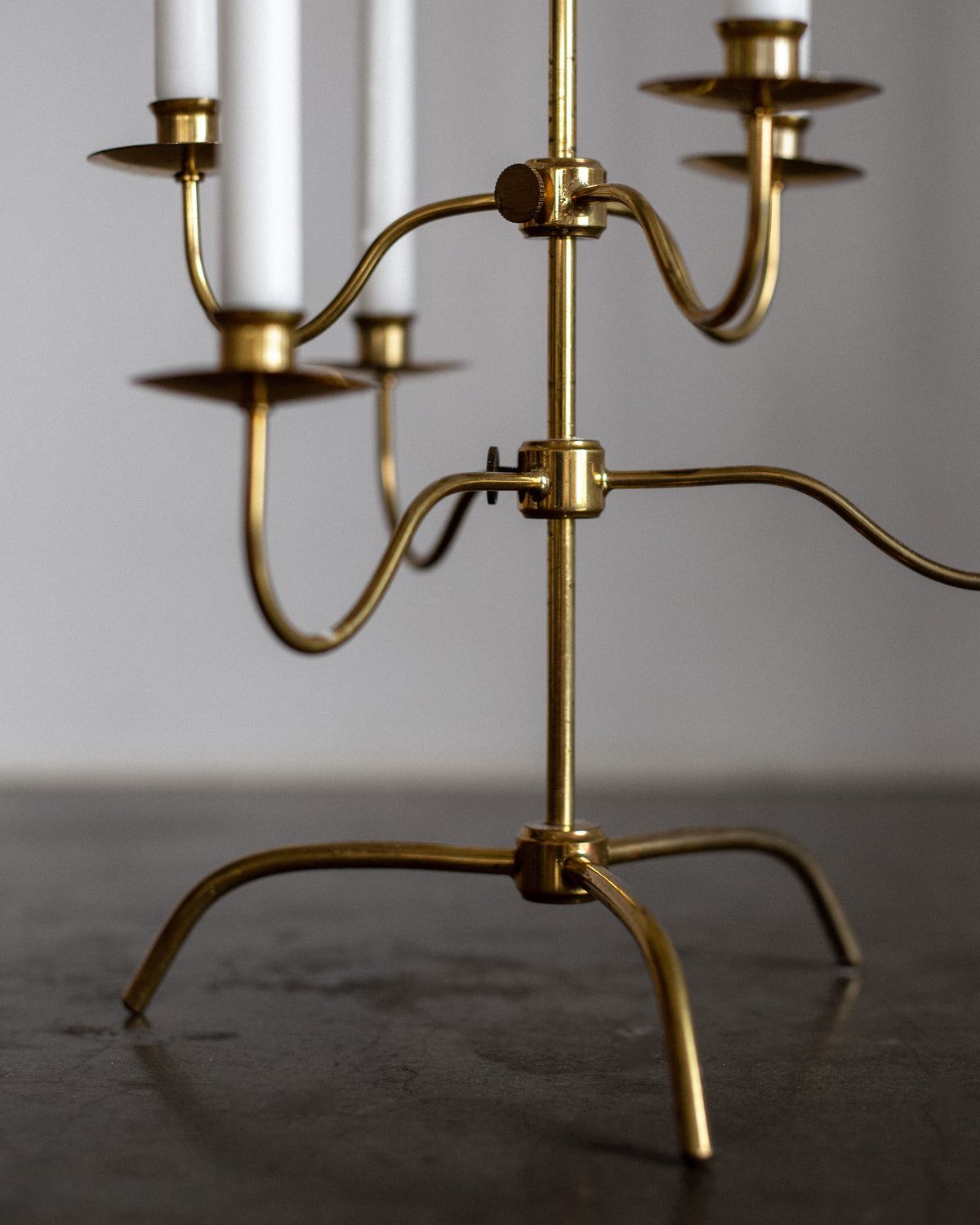 A close-up of a Spigel Swedish Mid-Century Brass Candelabra with four unlit white candles. Its elegant curved arms rest on a dark, smooth surface, while the softly blurred background beautifully emphasizes its intricate details.