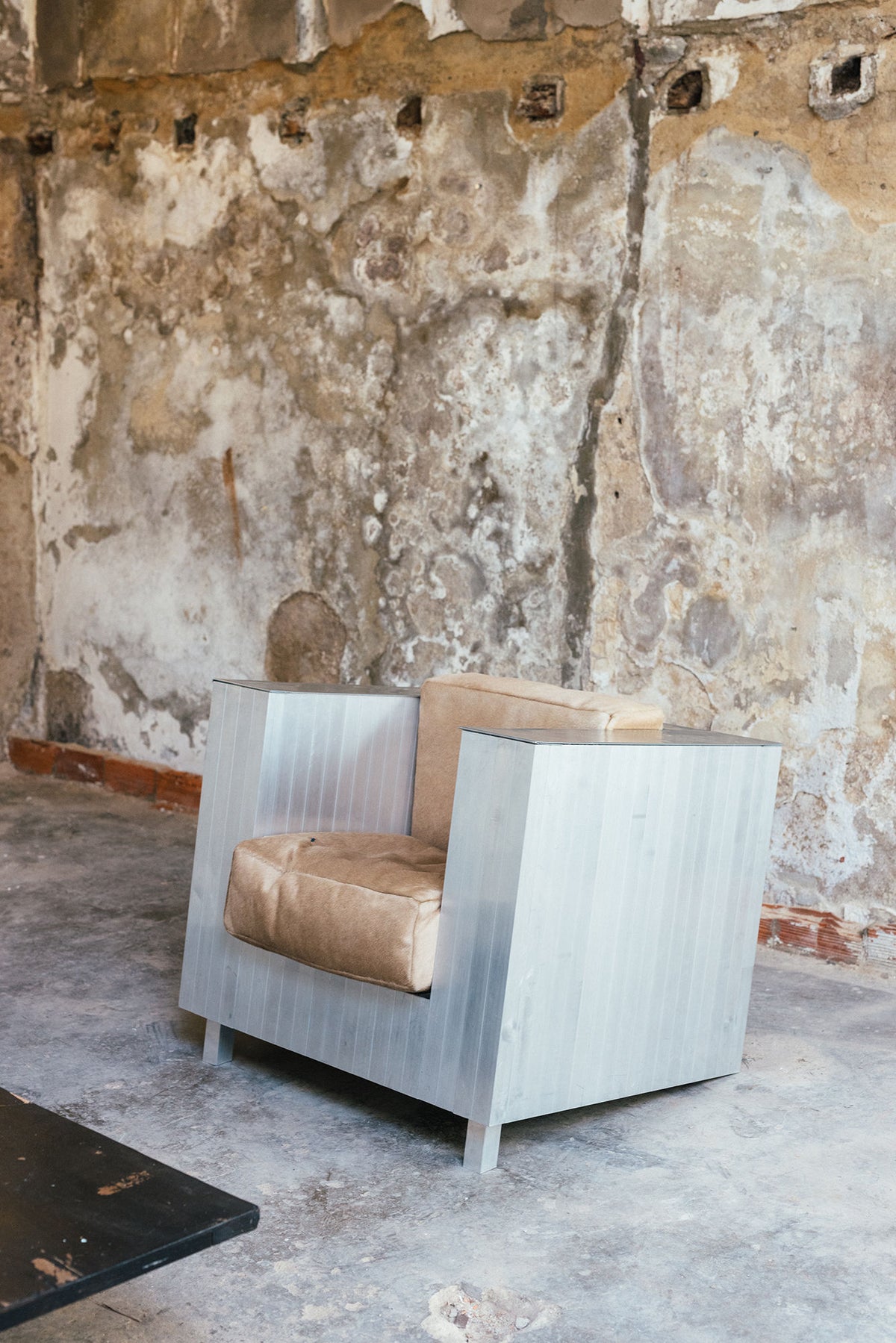The Aluminum Armchair by Studio HAOS, featuring tan cushions, stands out in a rustic room with weathered plaster walls and a concrete floor—showcasing the brand’s contemporary design style.