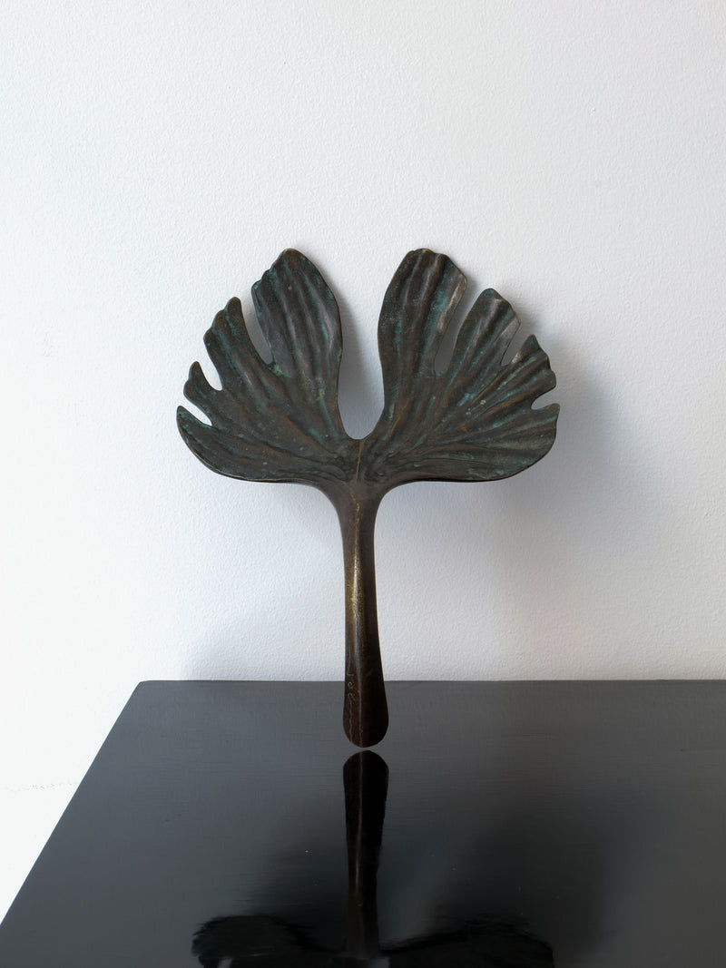 The Bronze Jobin Sculpture by Collection apart, designed to resemble a stylized ginkgo biloba leaf with two broad and textured lobes, is showcased on a glossy black surface against a white background.