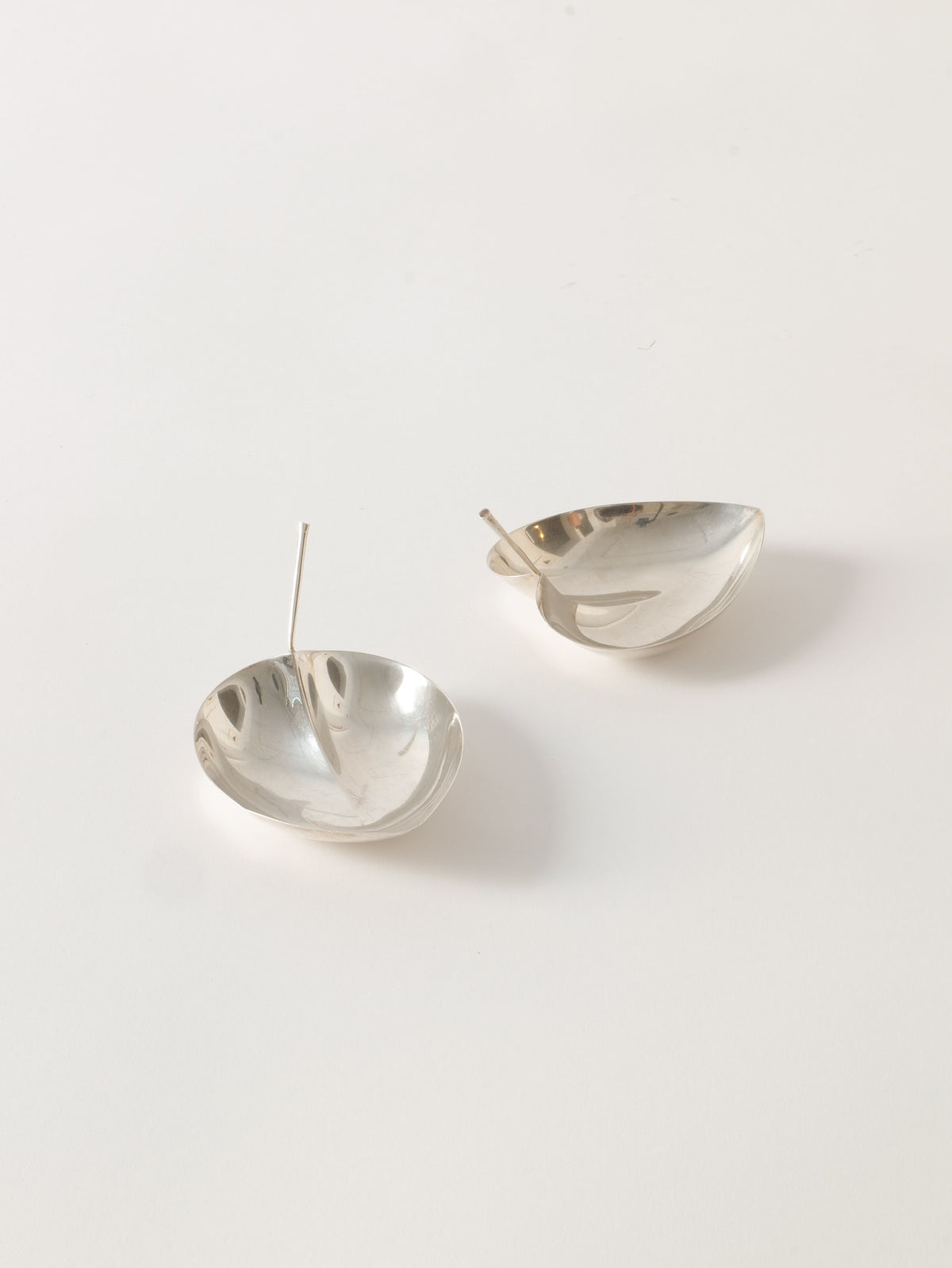 A pair of minimalist, Tapio Wirkkala Silver Bowls by Collection apart with a unique, shallow, concave design. Reminiscent of Tapio Wirkkala silver bowls, they are displayed on a white surface, each earring featuring a thin, straight post for fastening. The reflective surface showcases a sleek and modern aesthetic.
