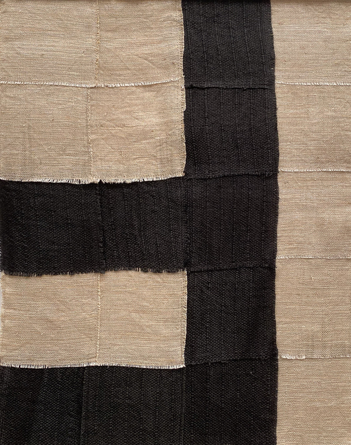The "Paths" pattern by Sundance Studio features large alternating light beige and black squares, crafted from antique cotton. With visible stitching forming a textured grid, this design exudes a rustic, handcrafted aesthetic.