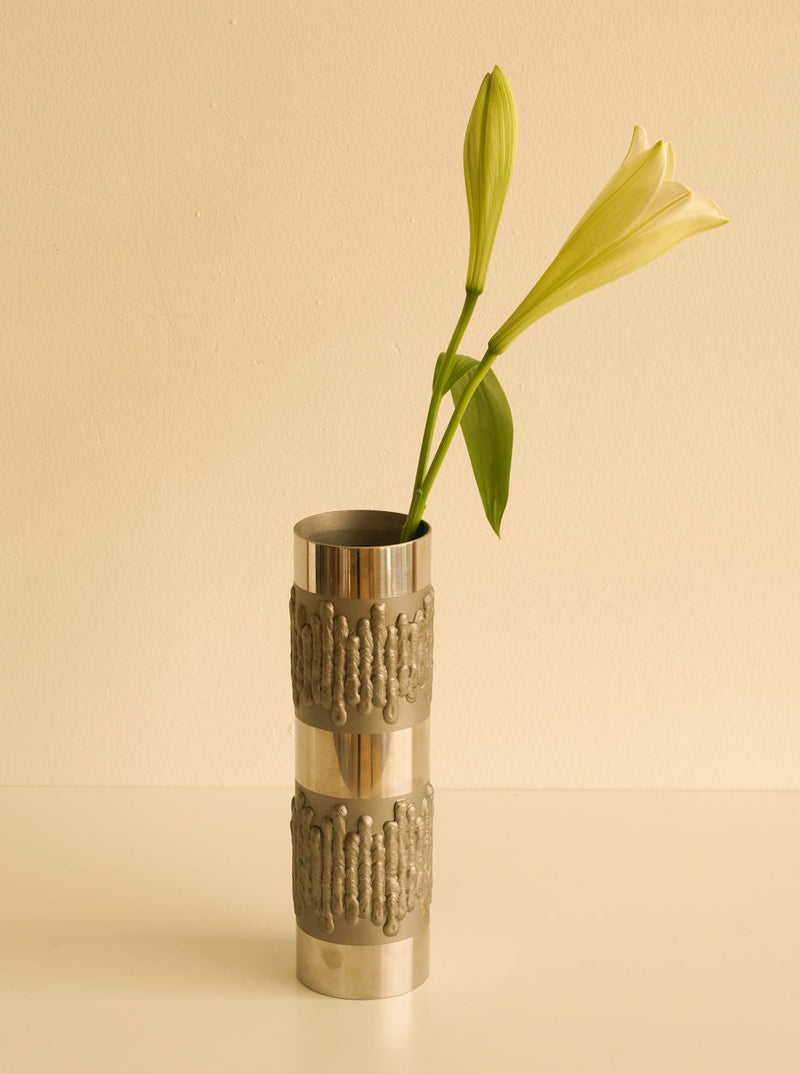 A single lily flower with buds and leaves is elegantly displayed in a mid-century brutalist Les Objets Amsterdam vase, placed against a light beige background.