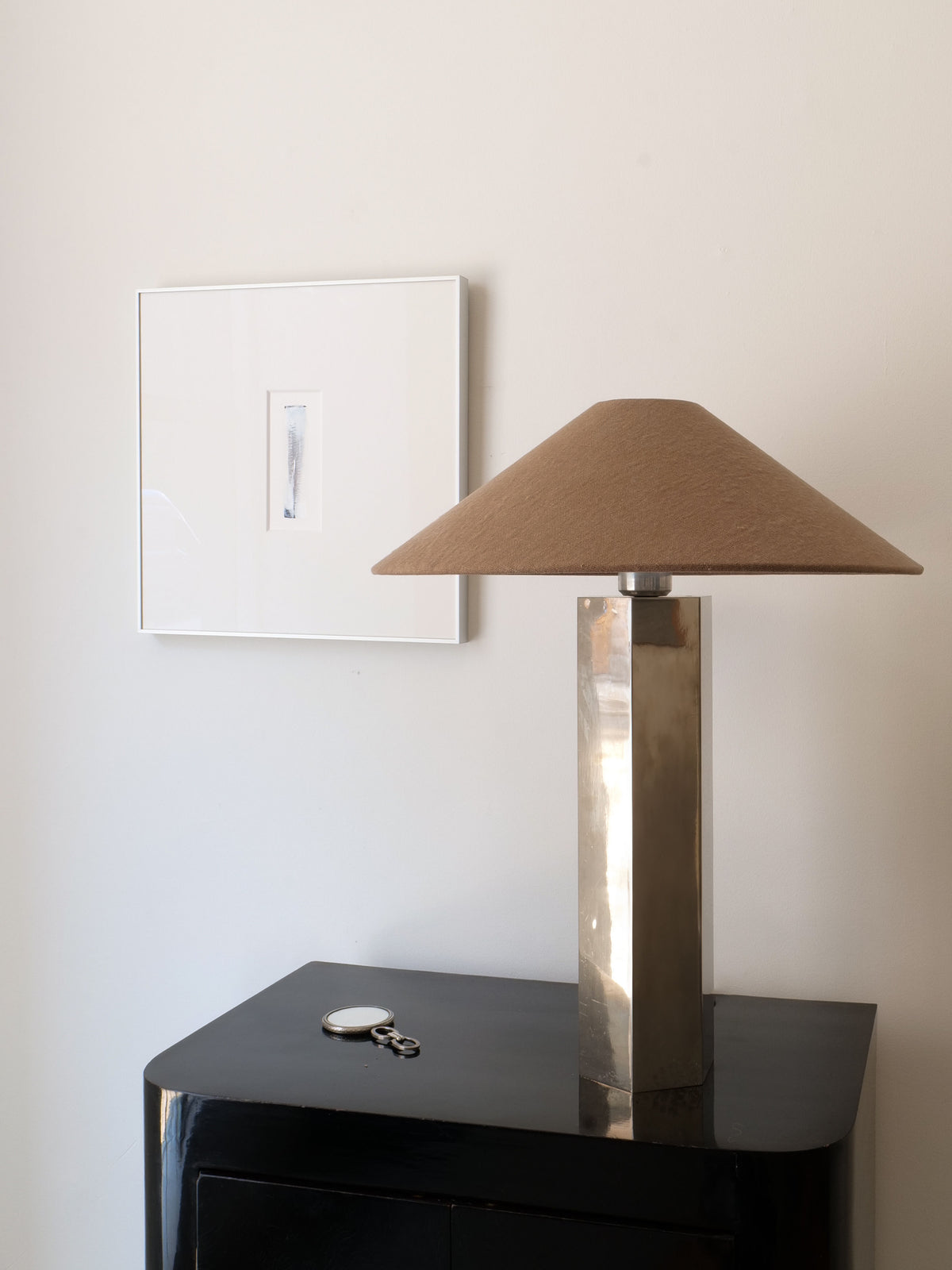 The Grand Hexagonal Lamp by Collection apart, featuring a brown lampshade, sits on a glossy black cabinet alongside round eyeglasses and a small white item, with minimal framed artwork on the light-colored wall behind.