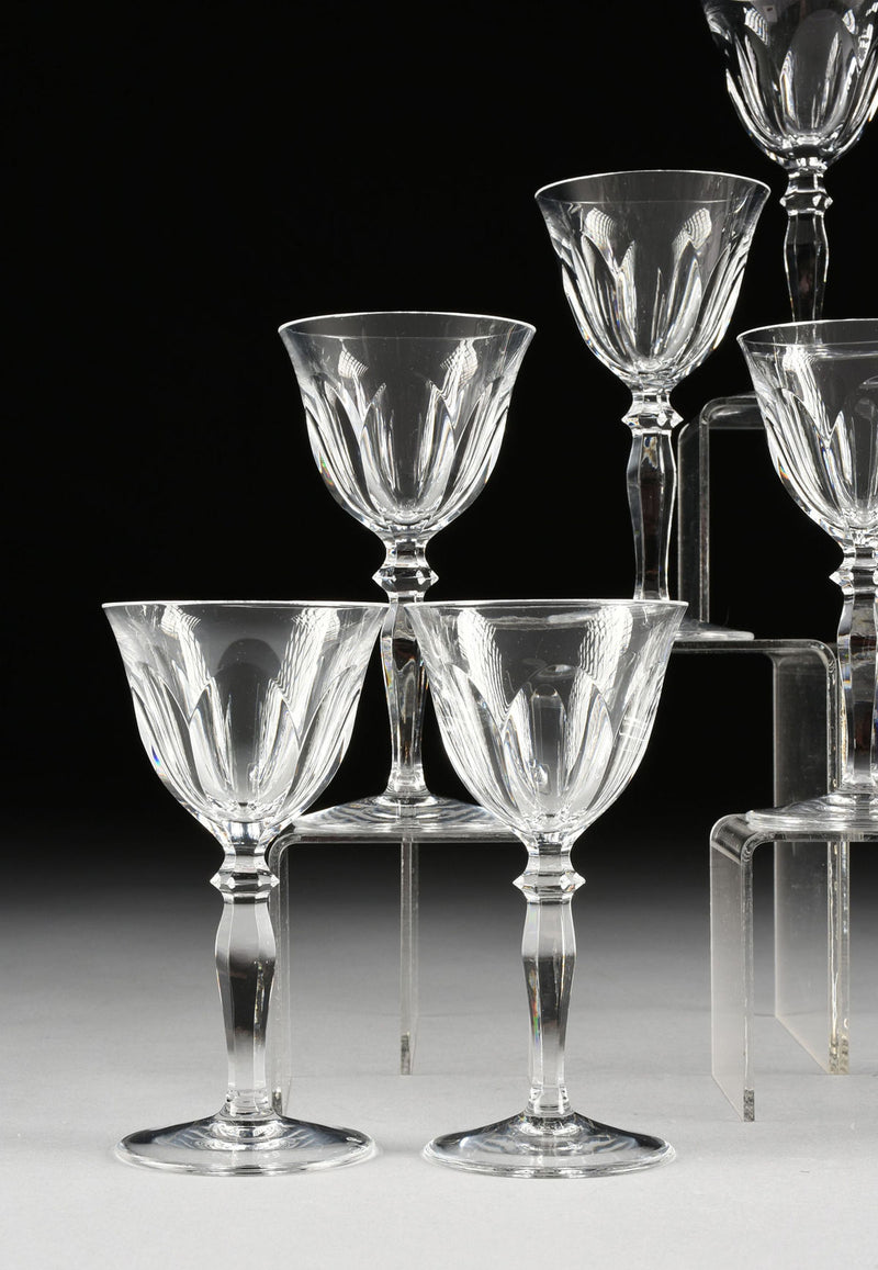 A set of 6 Peill & Putzer crystal wineglasses by Flock Studio, featuring faceted detailing and wide bowls, are displayed on clear risers against a dark background and light gray surface.