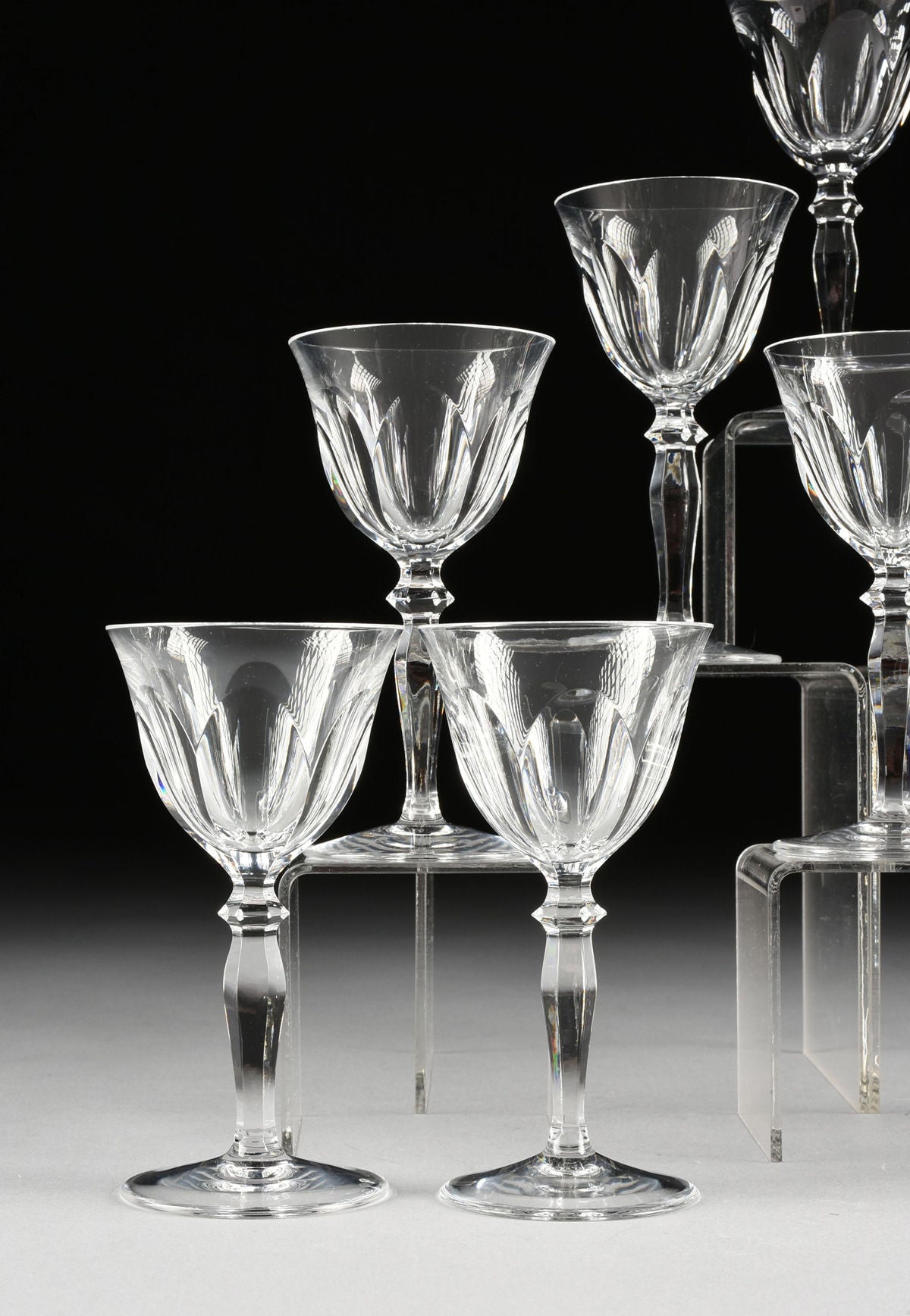 A set of 6 Peill & Putzer crystal wineglasses by Flock Studio, featuring faceted detailing and wide bowls, are displayed on clear risers against a dark background and light gray surface.
