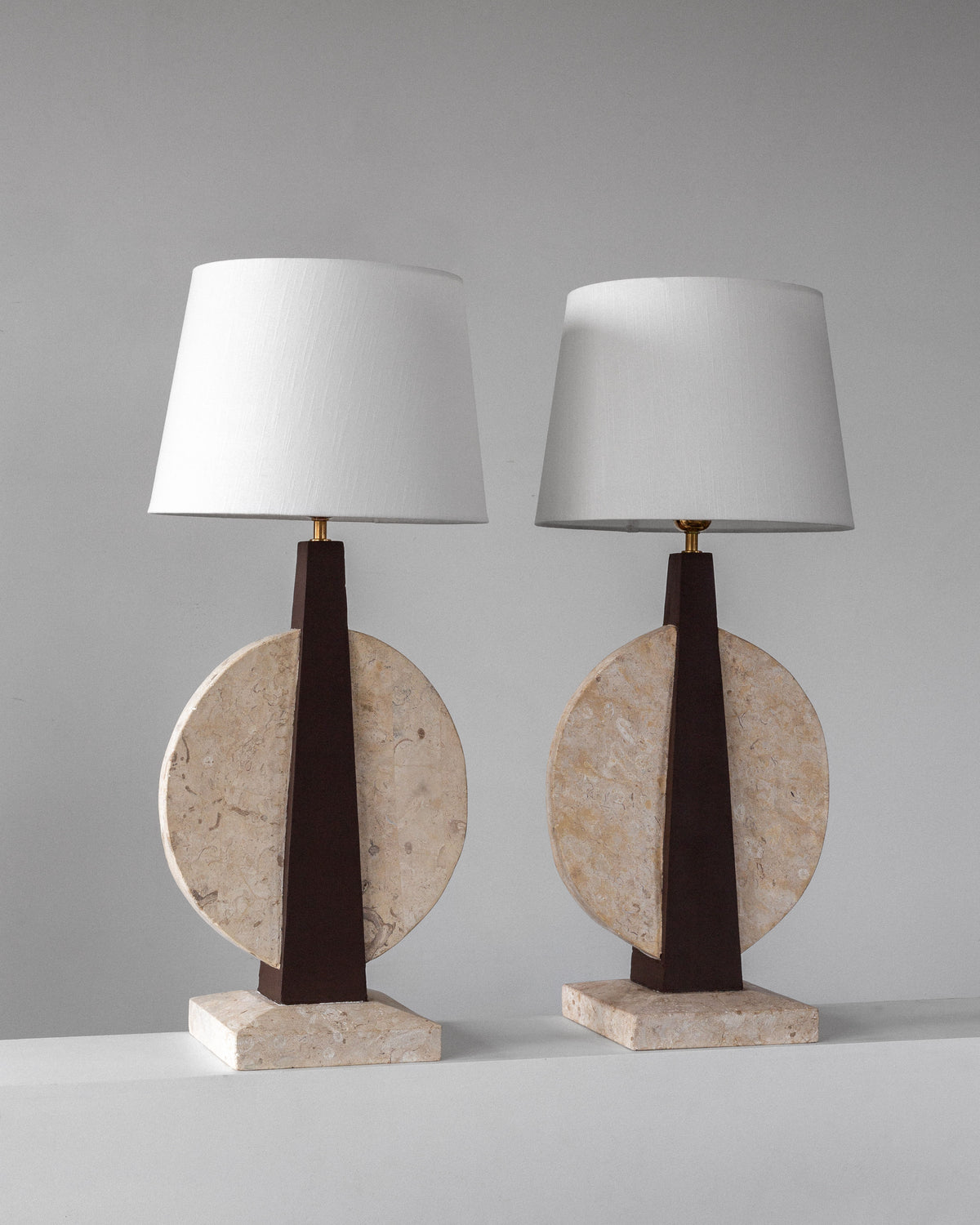 Spigel's Pair of Large Travertine Table Lamps from the 1970s feature modern white fabric shades with dark triangular stands and light-colored stone circular discs. These lamps rest elegantly on a white surface against a plain gray background, evoking vintage style.