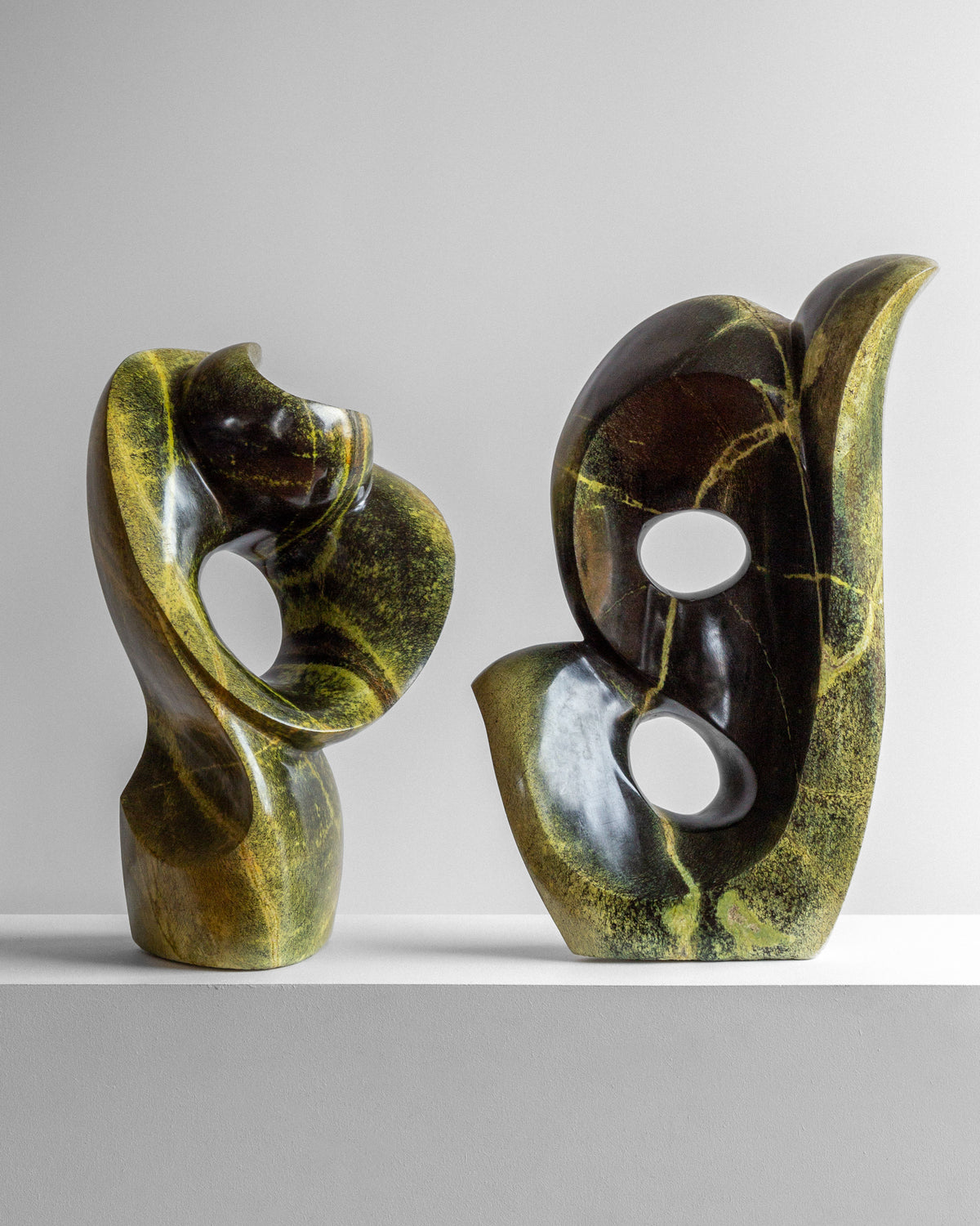Two "Abstract Stone Sculpture II" pieces by Spigel, crafted by a Zimbabwean artist, feature smooth curves and large cut-out holes. They rest on a light gray surface against a neutral backdrop, with a polished finish and modern design highlighting their striking abstract composition.
