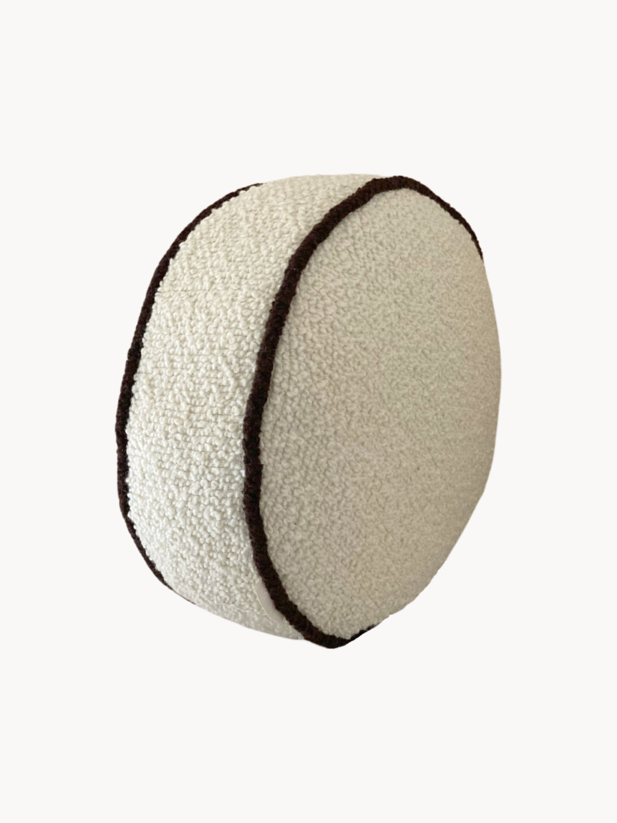 A round, plush cushion covered in white textured fabric, with parallel black lines running along its sides, resembling the stitching on a sports ball. This vintage-inspired design is crafted using British eco wool for a touch of classic elegance and sustainable comfort. The Le Rond - Passepoil - Wool Bouclé - Egg White/Chocolate by AMÉLIORÉ.