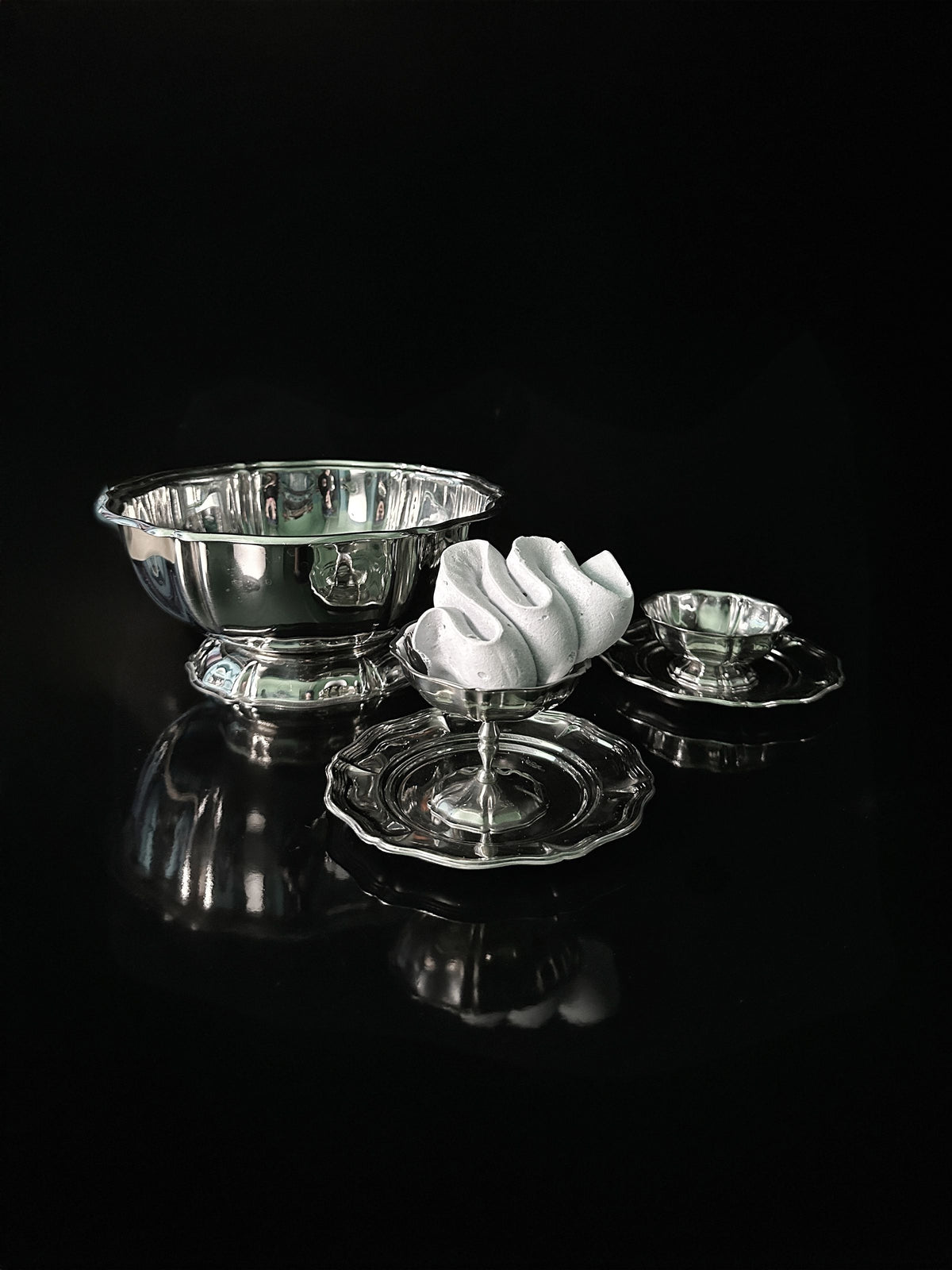 The Dodo Vintage Italian Stainless Steel Dessert Coupe Set, featuring a shiny silver bowl, smaller dish, and decorative glass stand with a white ornament, is elegantly arranged on a reflective black surface. The shimmering reflections add to its charm.