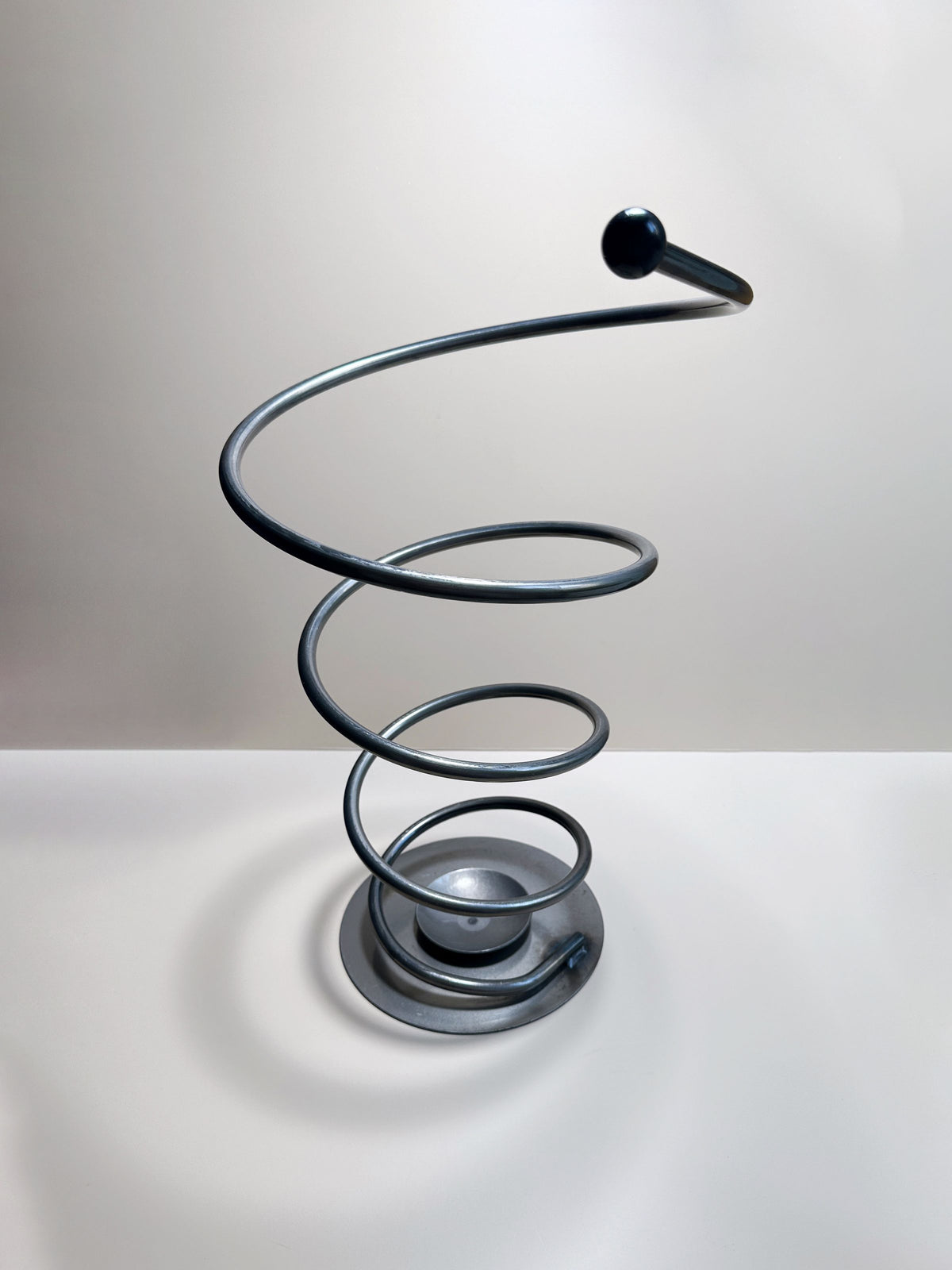 A chic Spiral Metal Umbrella Stand by Dodo Vintage showcases a post-modern design with a striking spiral shape and is crowned with a black sphere. It tapers elegantly from a wide circular base to a narrow end and is crafted from solid metal. This sculpture is artfully placed on a smooth, neutral-colored surface under soft lighting that enhances its modern aesthetic.