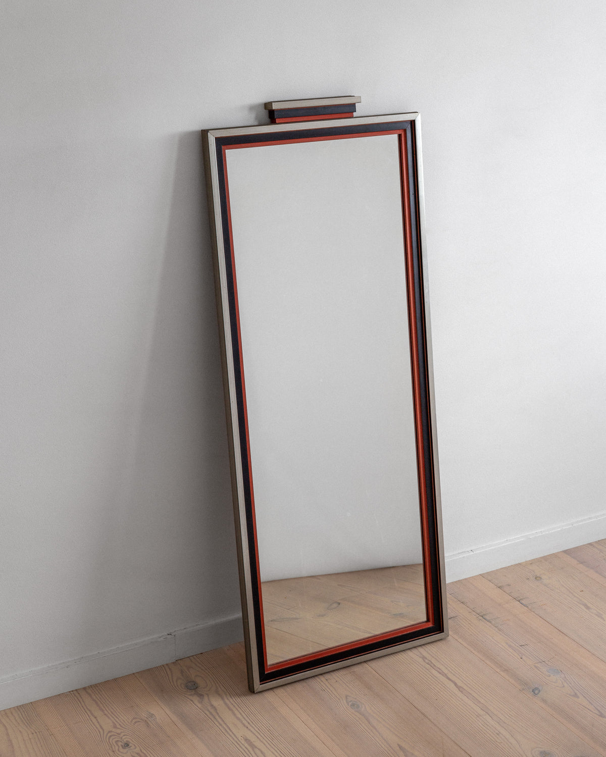 A tall, rectangular Art Deco Mirror from Spigel, dating back to 1930s Sweden, features a thin red border and a wider silver frame. It leans against a light gray wall on a wood floor with visible grain patterns, capturing J.A. Edenholm's timeless design essence.