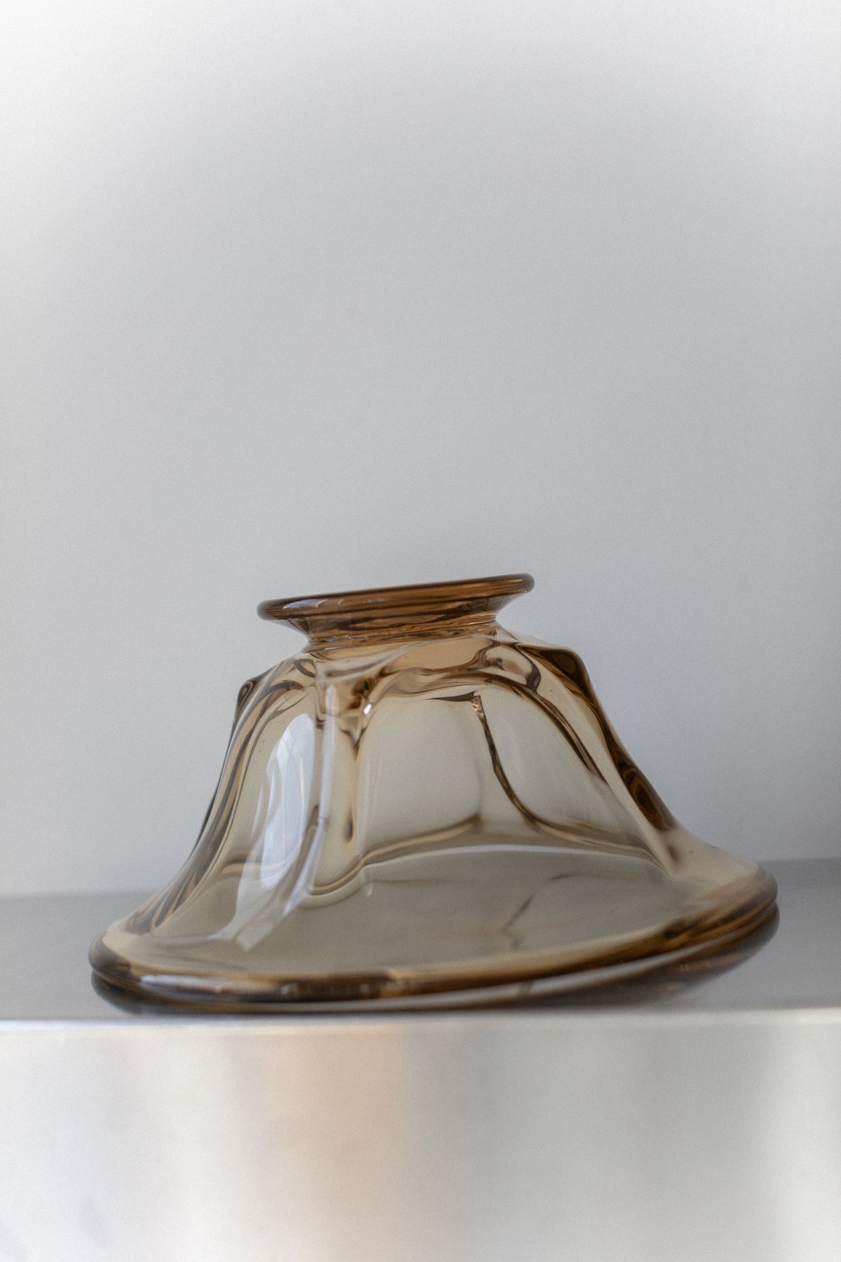 A vintage glass bowl from Out For Lunch, featuring a unique wavy shape in translucent brown with hints of yellow, is placed upside down on a smooth, light-colored surface. Its form resembles flowing liquid, contrasting beautifully with the minimalist background and evoking mid-century design aesthetics.