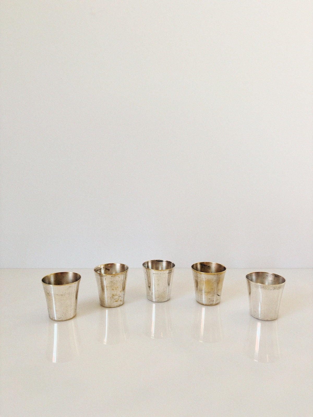 Five small, tarnished silver cups from the 60's Liquor Server Set by Imago Artis (Huerto) are evenly spaced in a row on a glossy white surface with a plain white background.